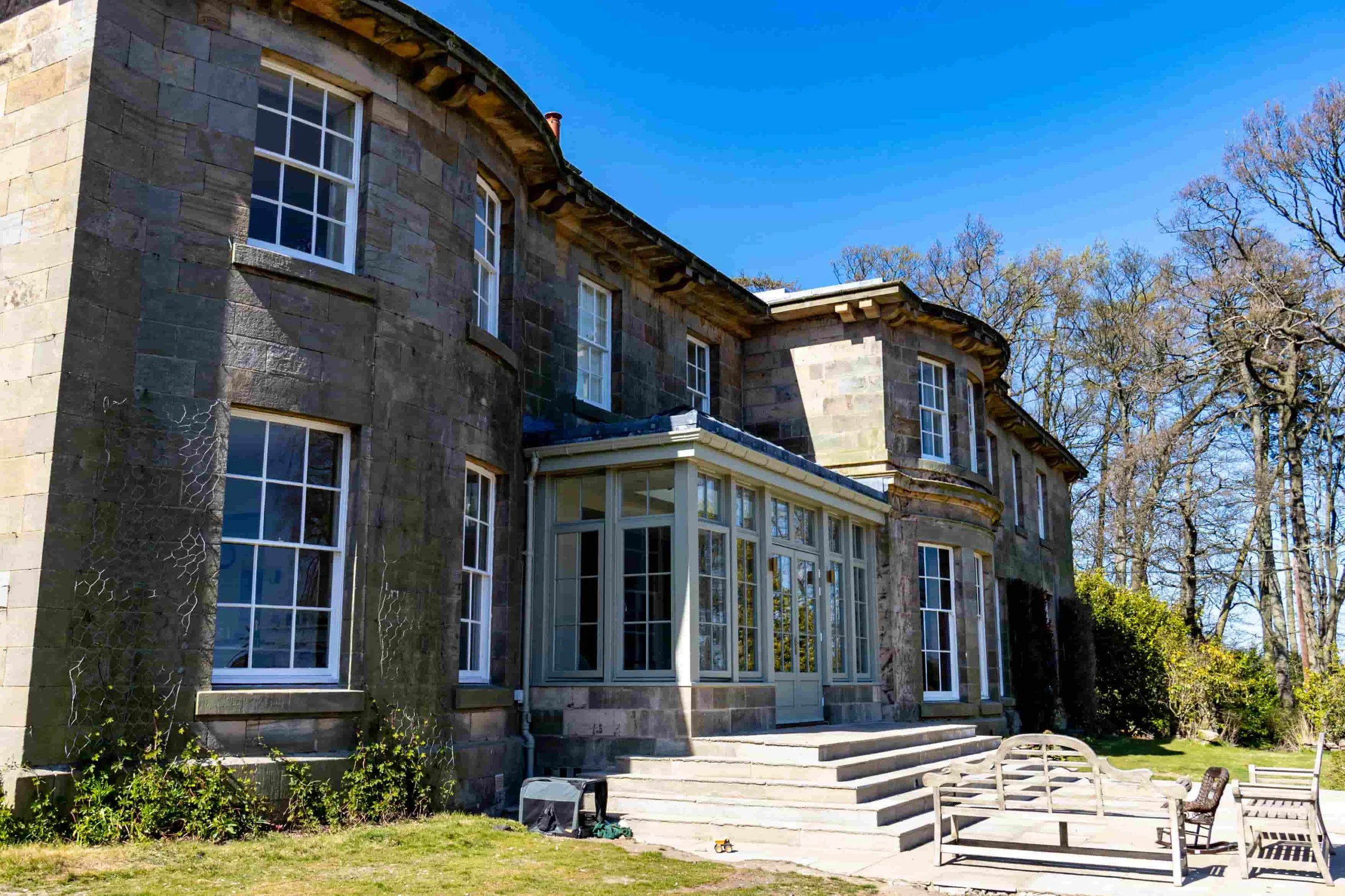 Back view of a large stone house with multiple white-framed windows, a glass sunroom, several wooden benches, and a grassy yard with tall leafless trees against a bright blue sky.