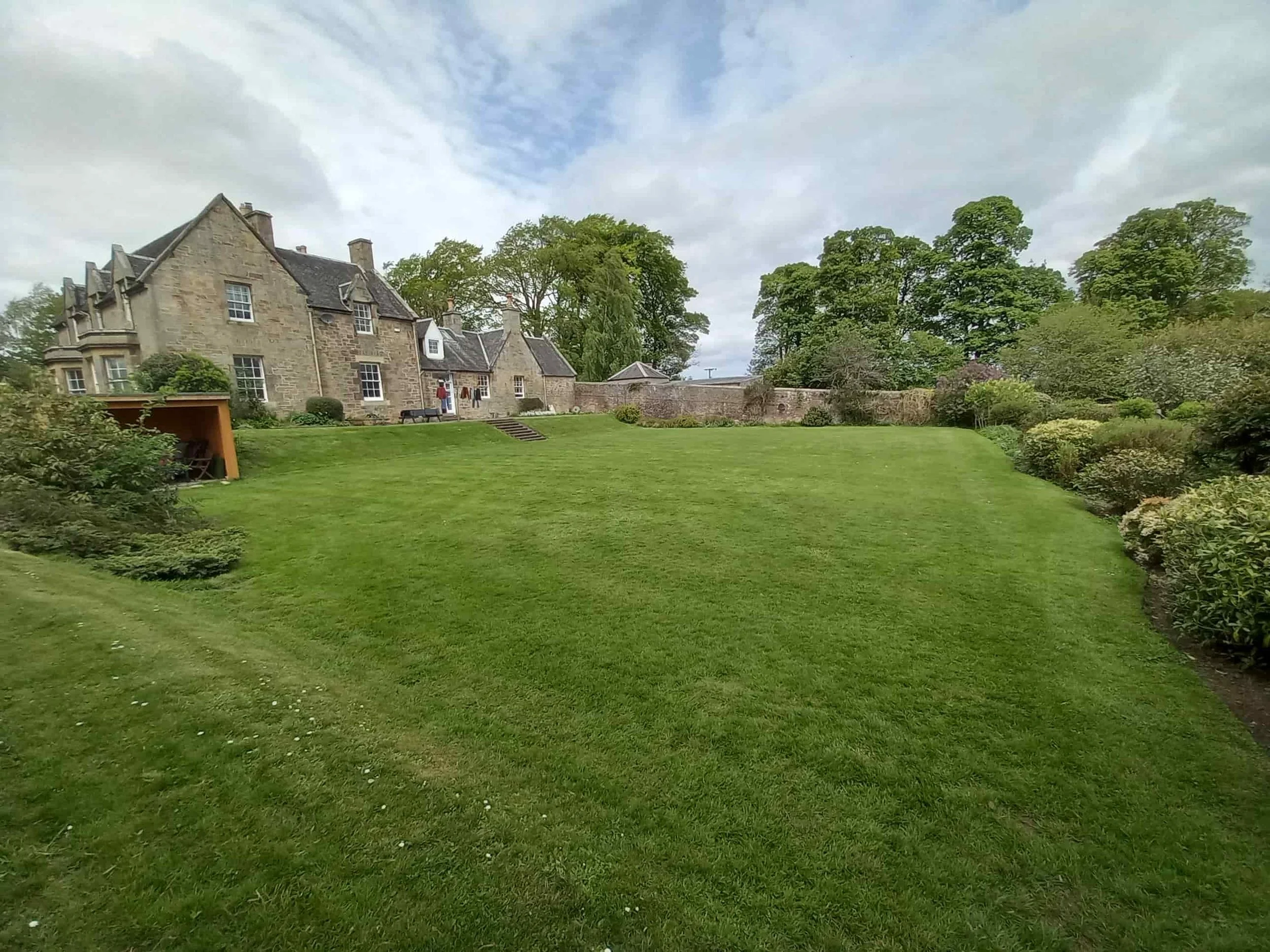 A large, well-maintained lawn in front of a historic stone house with multiple gabled roofs, surrounded by lush greenery and trees under a partly cloudy sky.