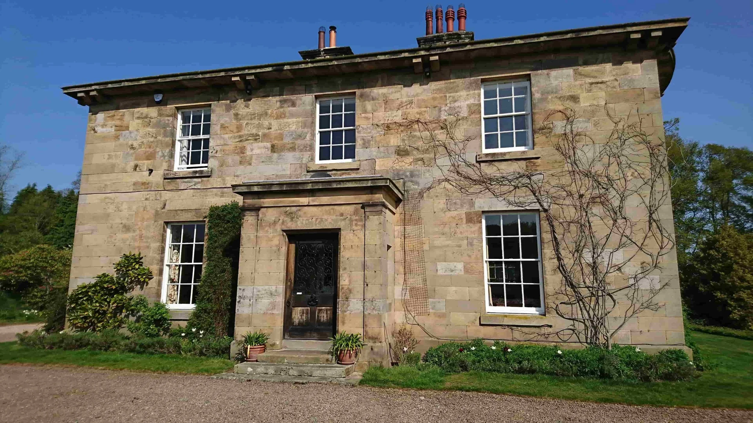A two-story stone house with five windows, a black front door, and climbing vines on the side, surrounded by a garden.