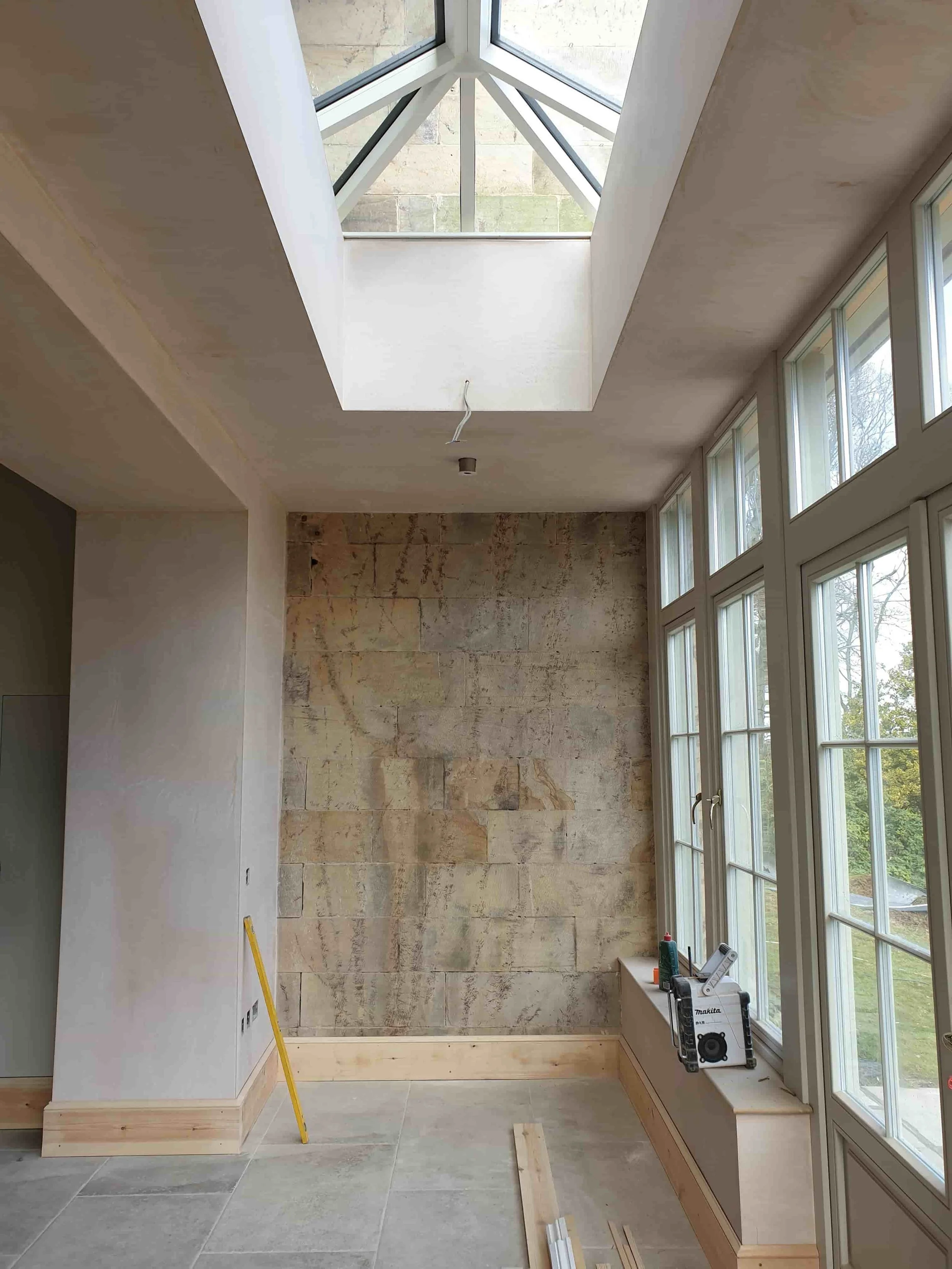 Interior view of a room under construction with large windows on the right, a partially finished wall with stone tiles, and a skylight overhead.
