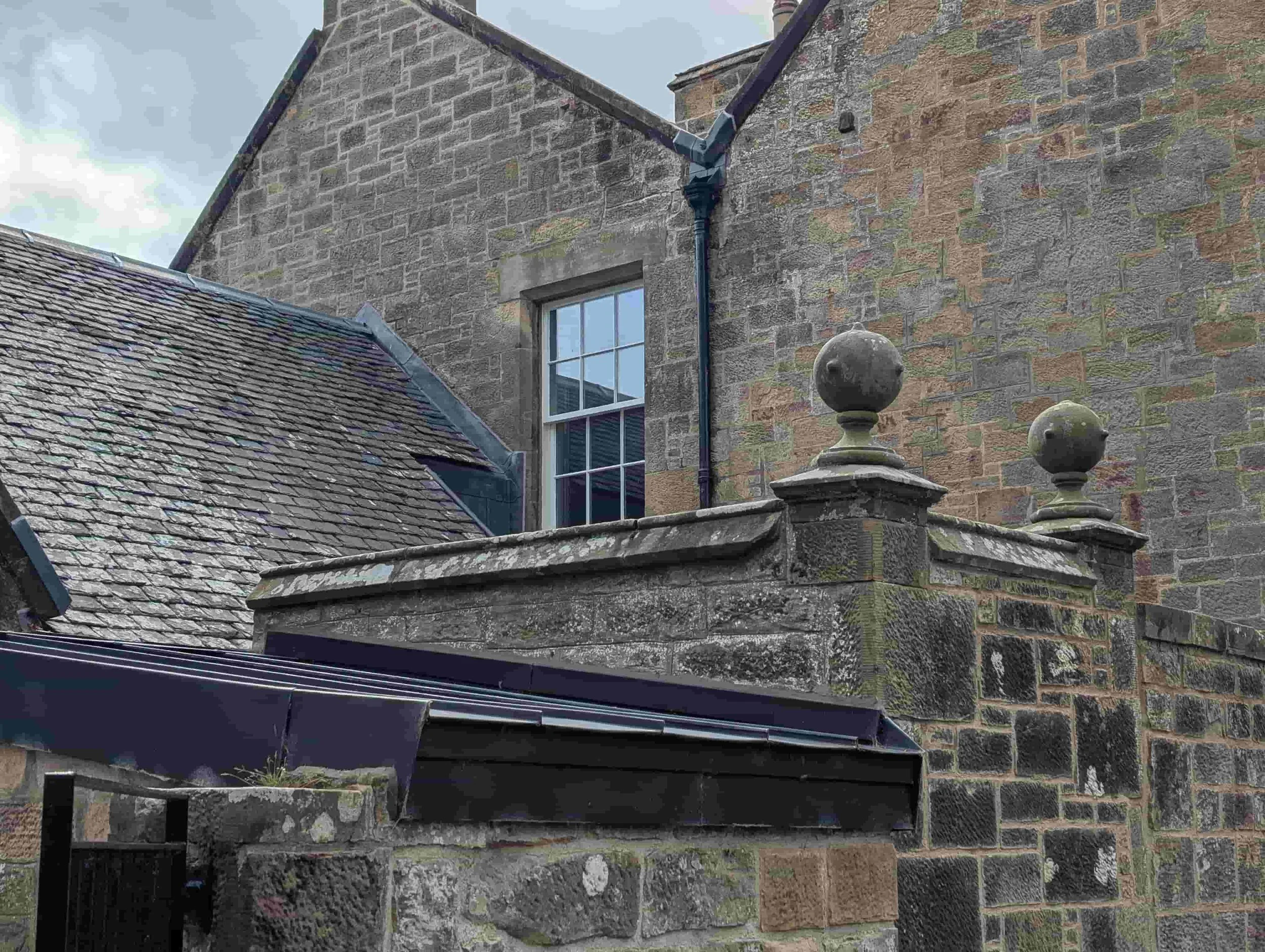 Close-up of an old stone wall with decorative stone balls on top, a window, and roofing structures, part of a historic brick building.