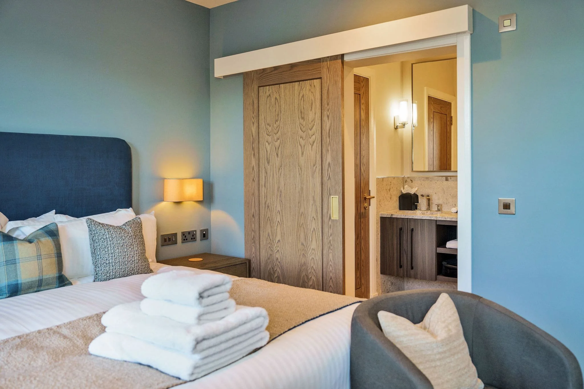 Clean and modern hotel bedroom with neatly made bed with pillows and a folded towel on top, soft lighting, and an open sliding door leading to a bathroom with a sink, mirror, and warm lighting.