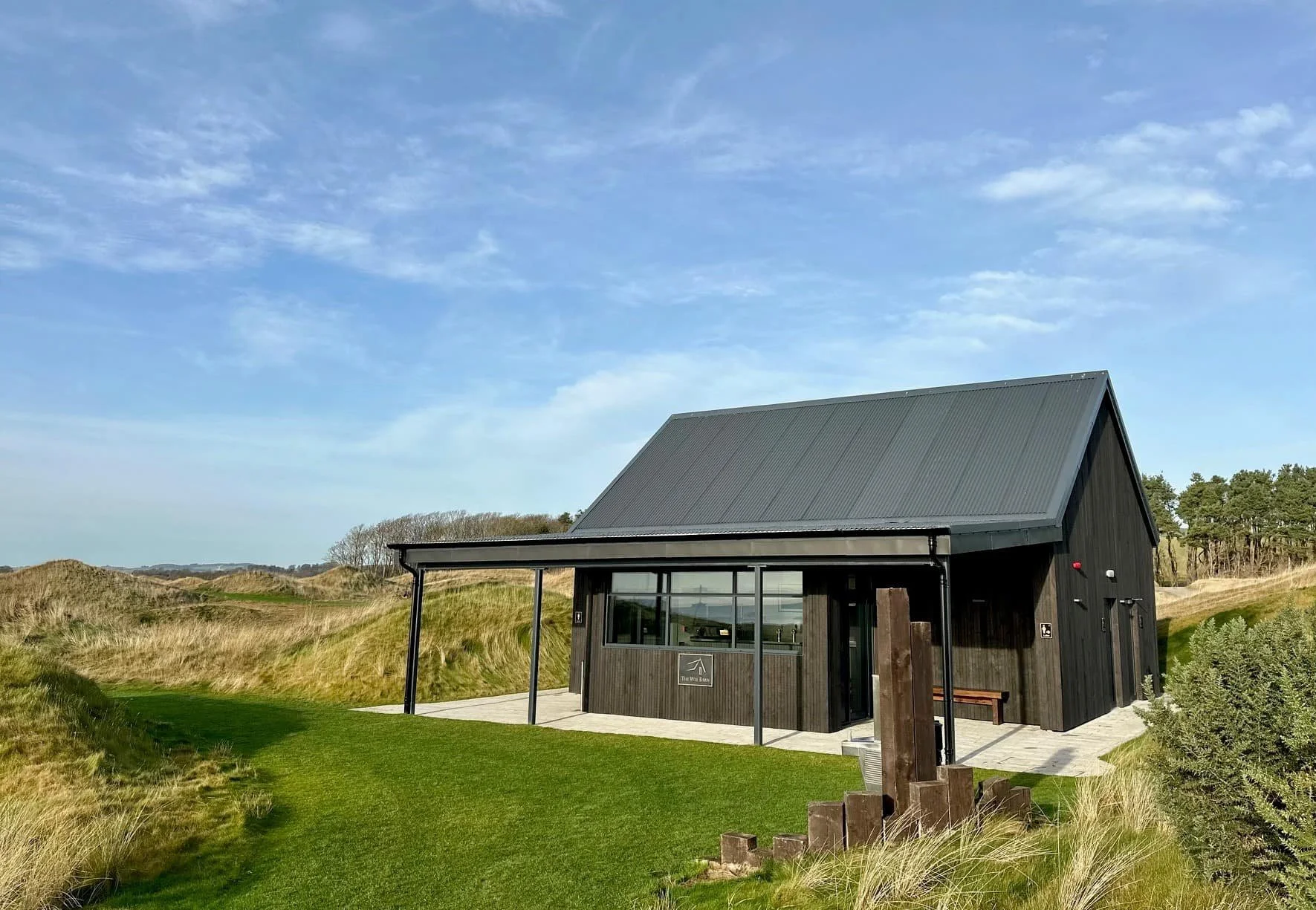 A black modern cabin with a sloped roof, large window, and a small patio, situated in a grassy landscape with sand dunes and sparse trees under a partly cloudy blue sky.