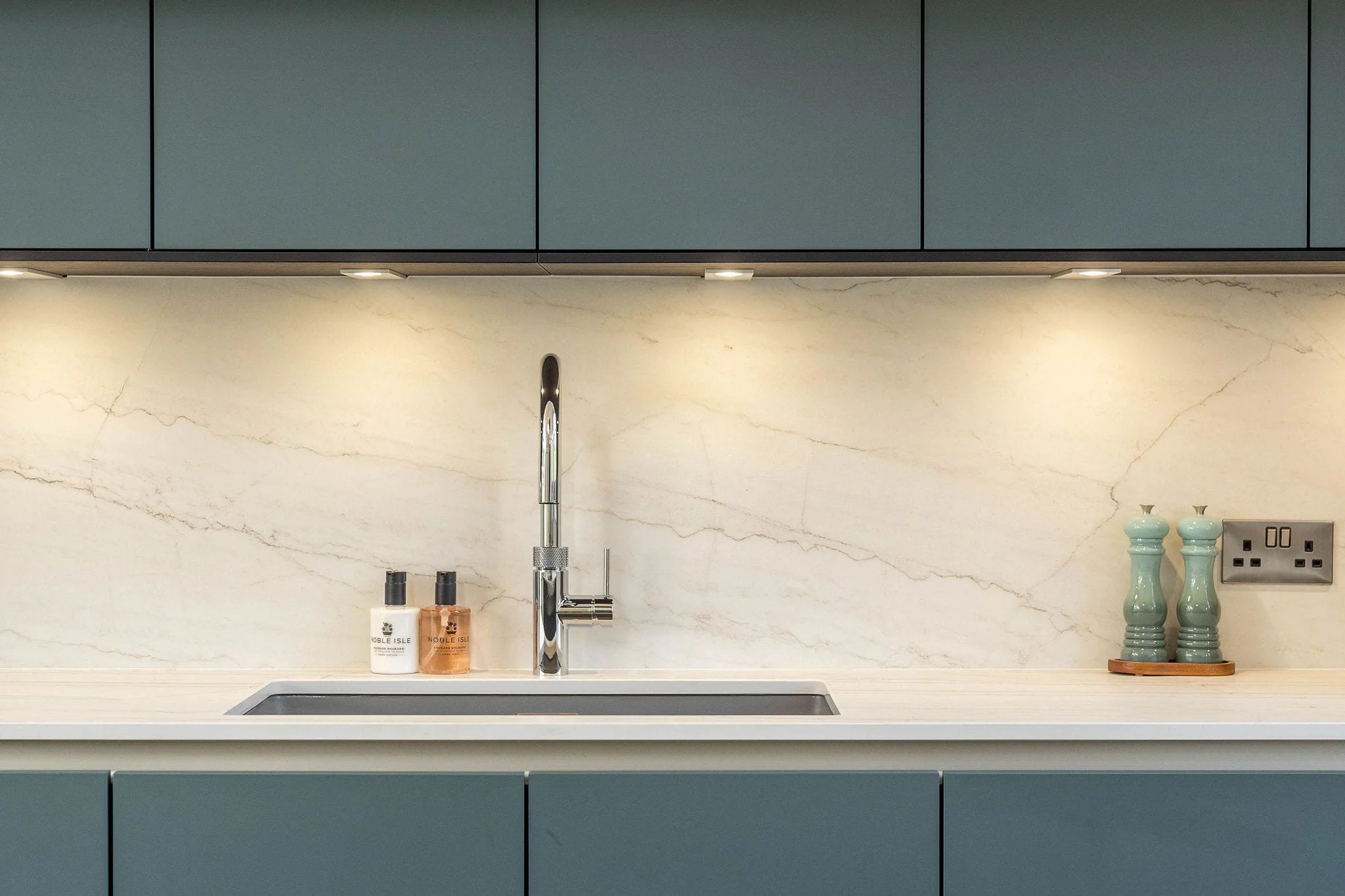 Modern kitchen with a marble backsplash, a stainless steel faucet, and blue cabinets. Items on the counter include soap dispensers, decorative vases, and a power outlet.