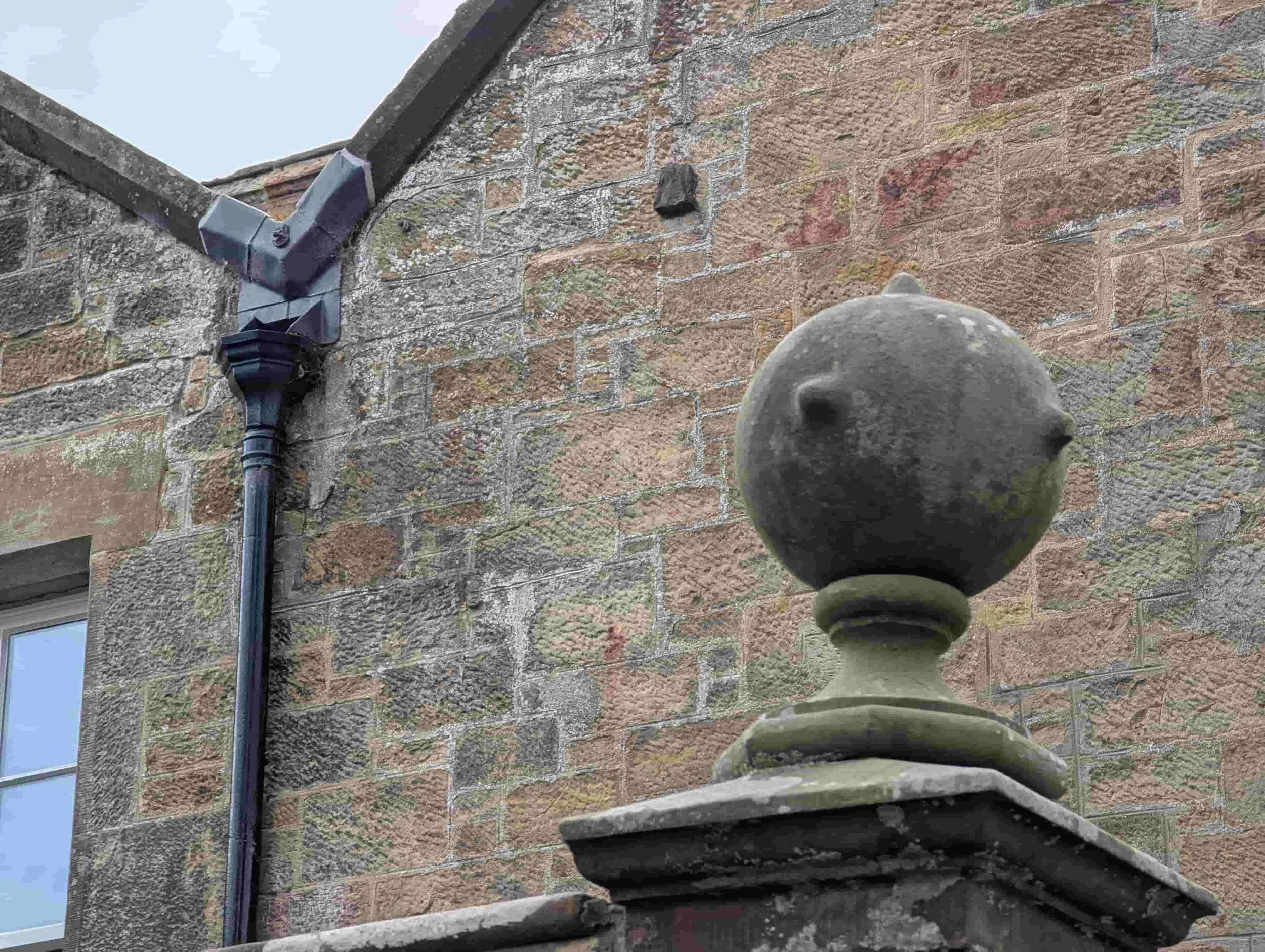 Close-up of an old metal finial on a stone wall in front of a brick building with a window, a gutter, and a drainpipe.