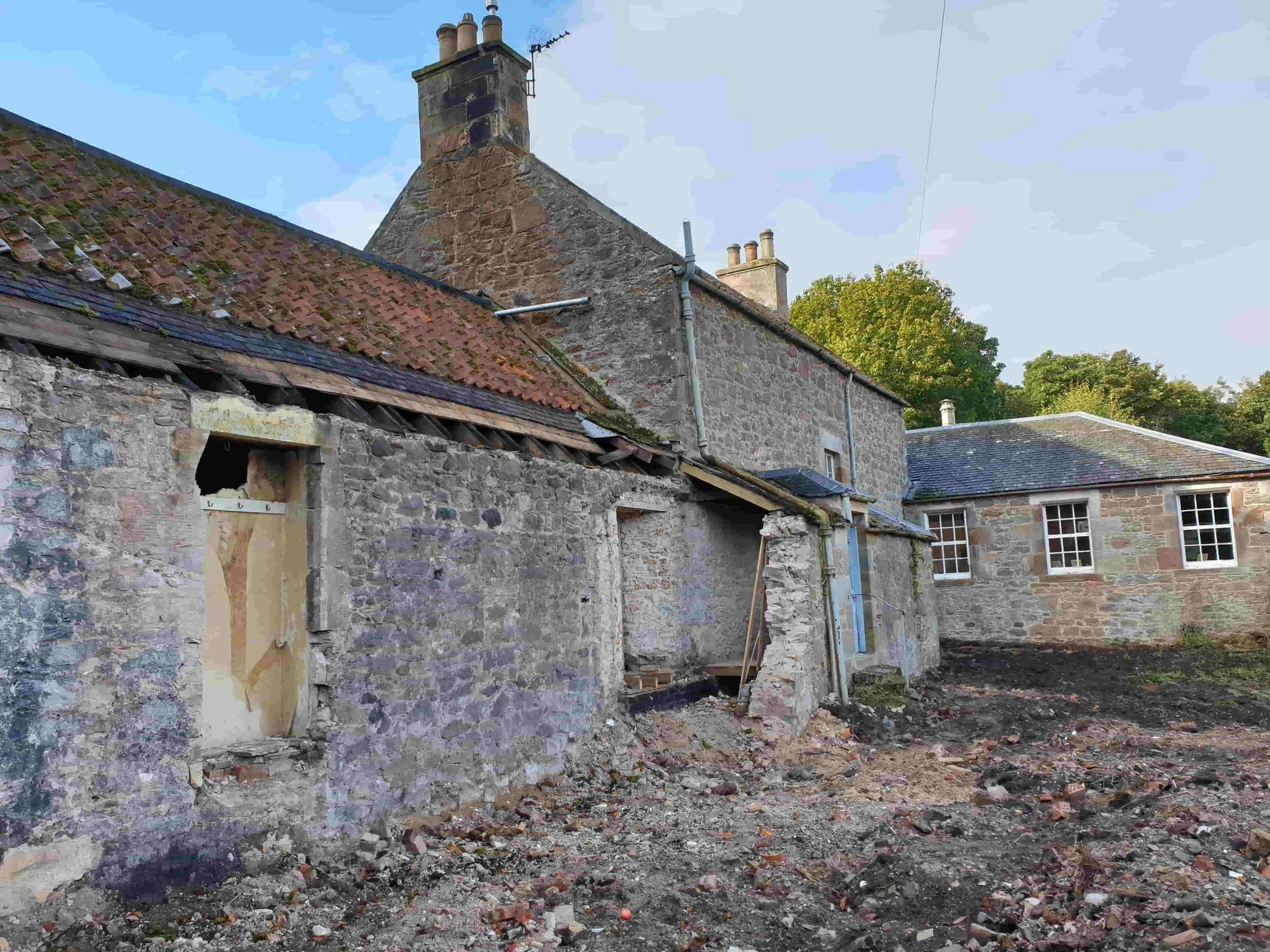Old stone house with renovation work in progress, missing window on the front wall, construction debris and exposed bricks on the ground, surrounded by other stone buildings, trees in the background.