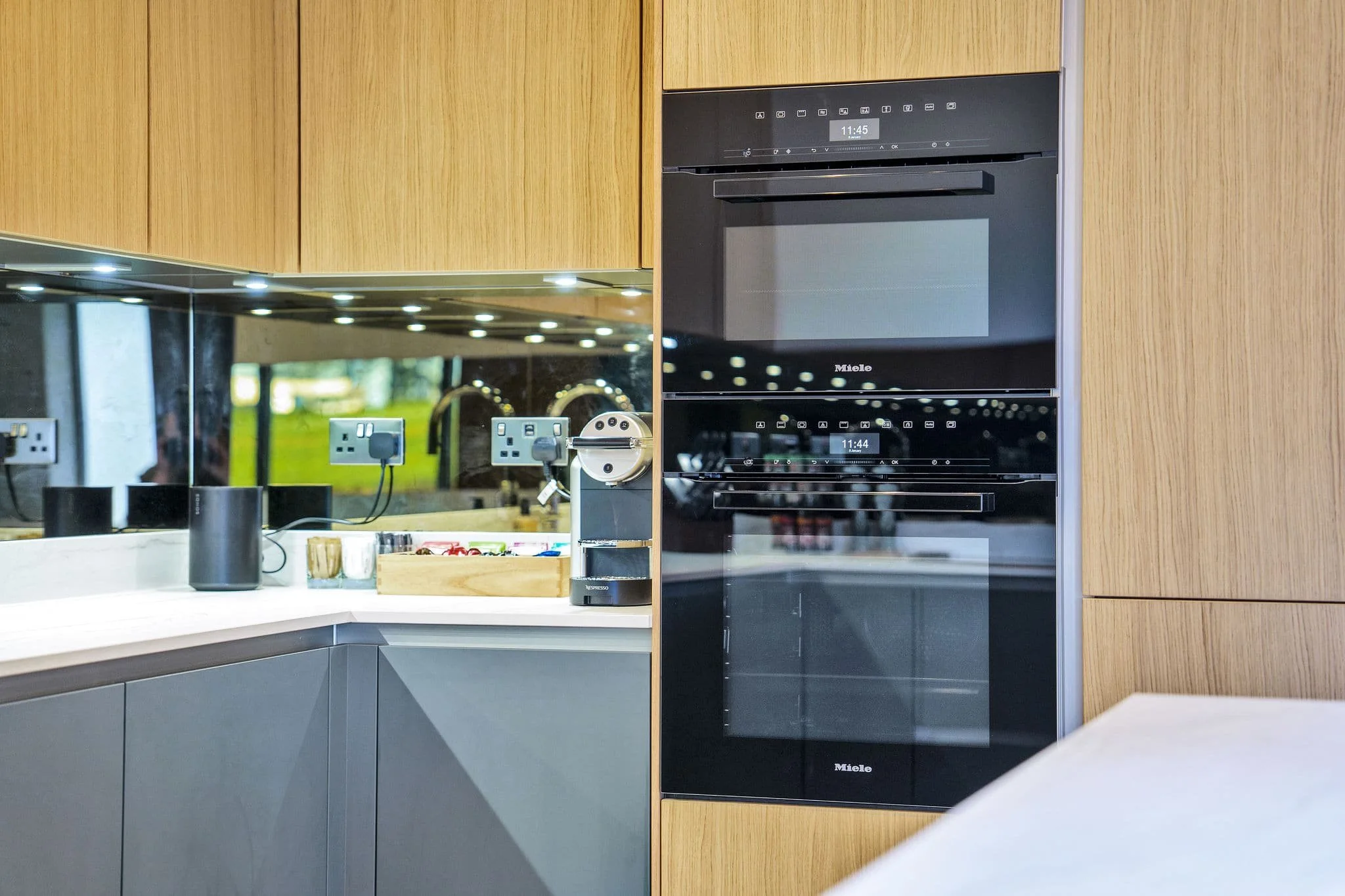 Modern kitchen with black built-in oven and microwave, wooden cabinets, white countertop, and a reflective backsplash.