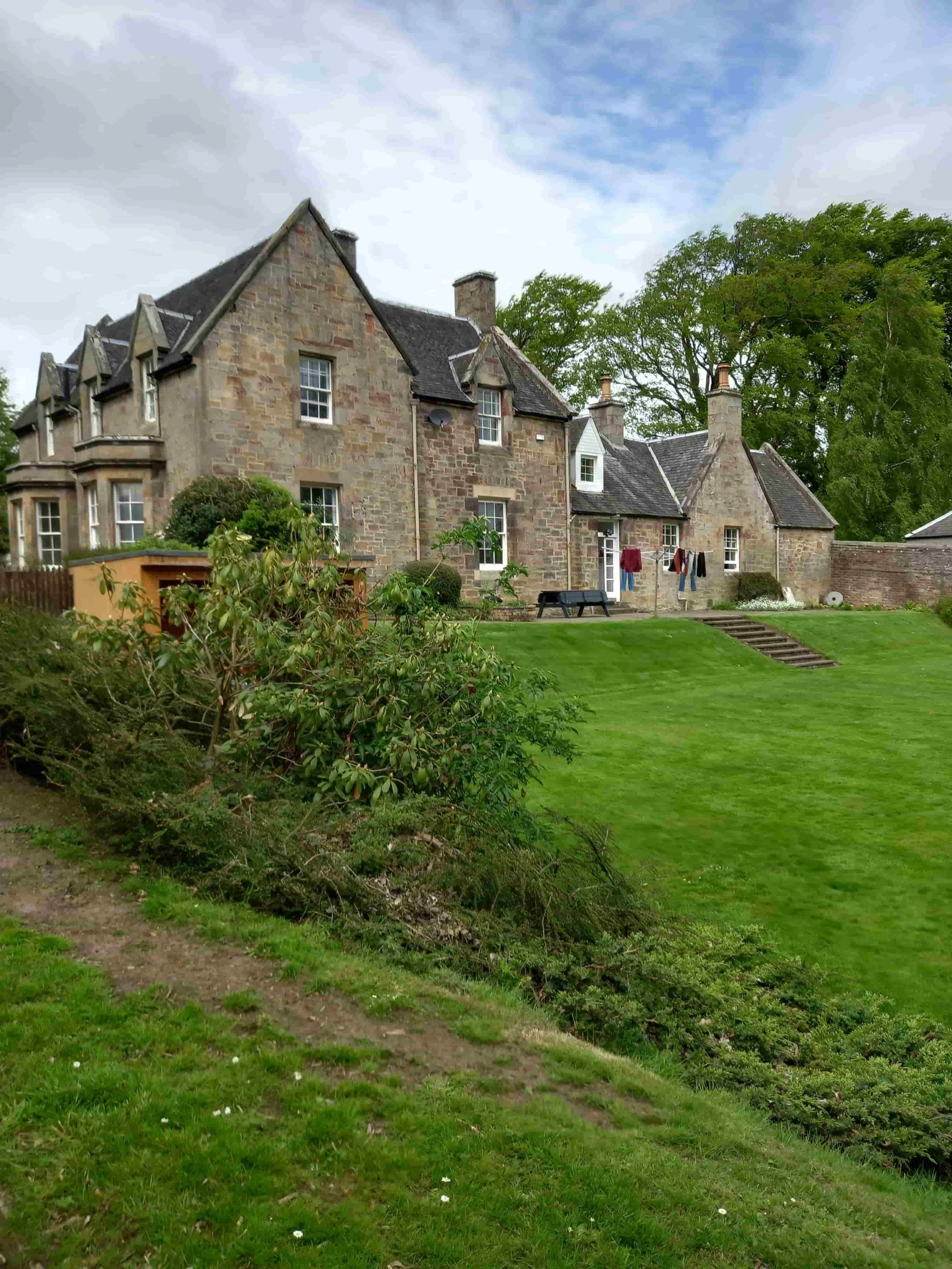 A large stone house with multiple chimneys, surrounded by green trees and a well-maintained lawn. Clothes are hanging outside to dry, and there is a black bench on the yard.