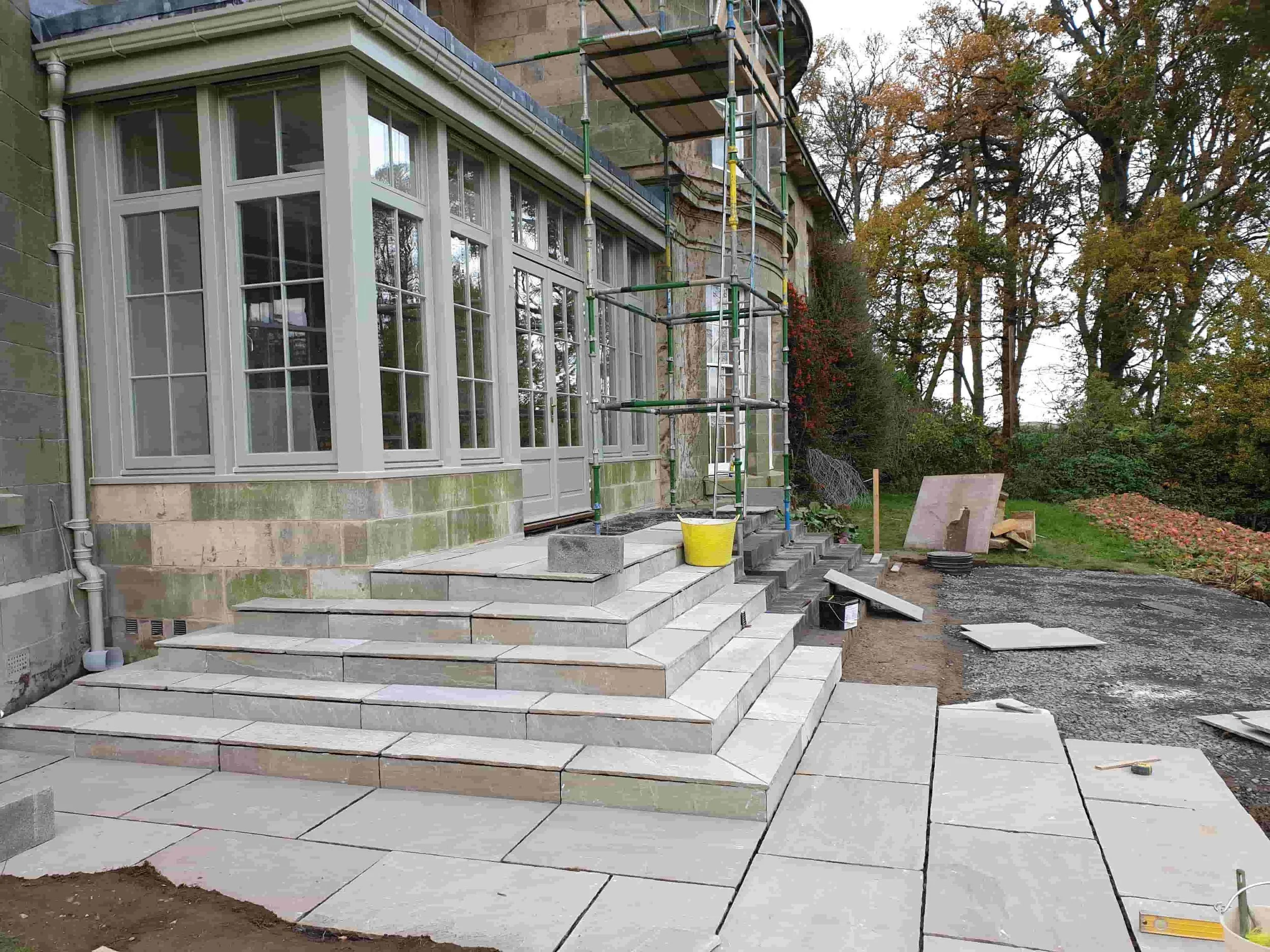 Construction site with staircase and scaffolding in front of a house with large bay windows, surrounded by trees and fall foliage.