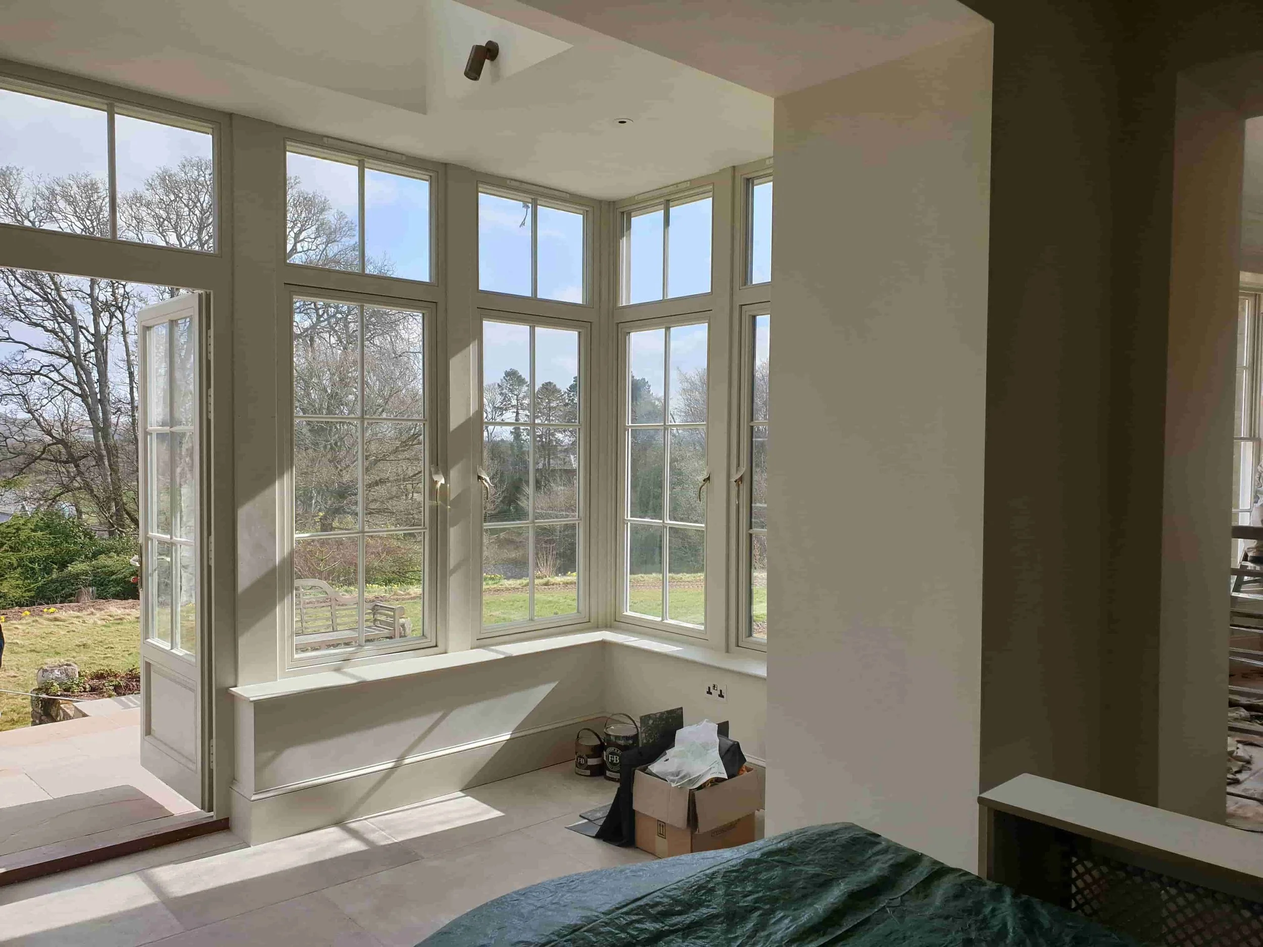 Bright indoor room with large bay window overlooking a garden with trees and a bench, construction supplies and a cardboard box near the window, and part of a bed with a dark green covering.