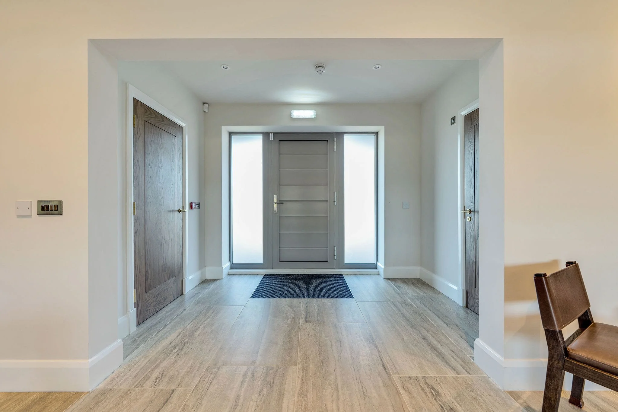 Modern entryway with wooden double doors, side glass panels, and a central front door with a handle, all illuminated by ceiling lights.