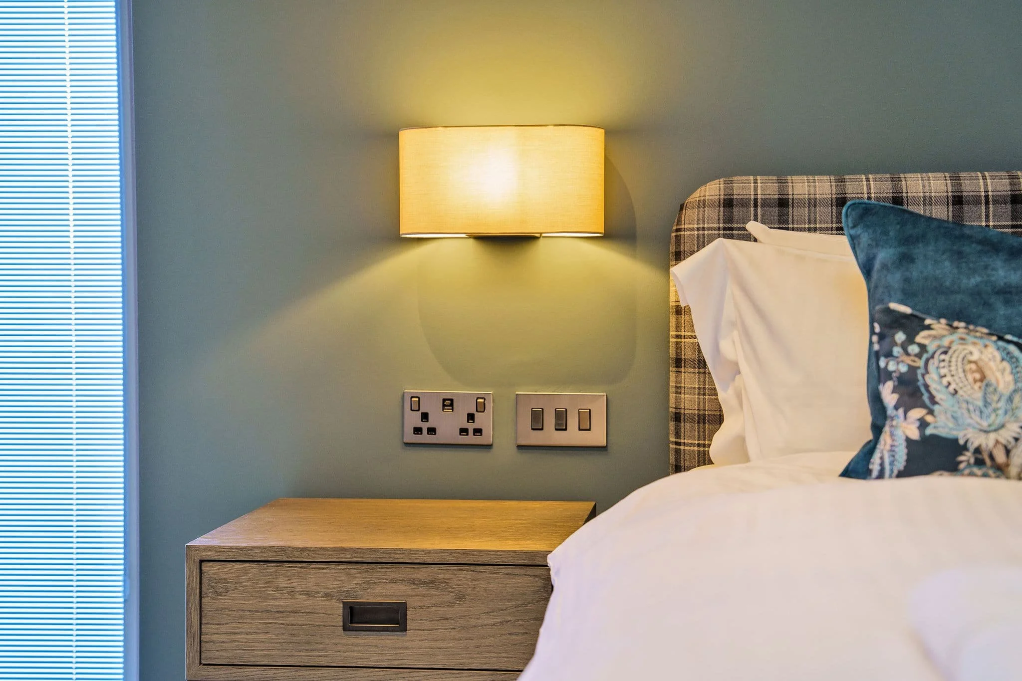 Close-up of a bedroom bedside table with a wall-mounted lamp, power outlets, and light switches; part of a bed with pillows and a checkered headboard is visible.