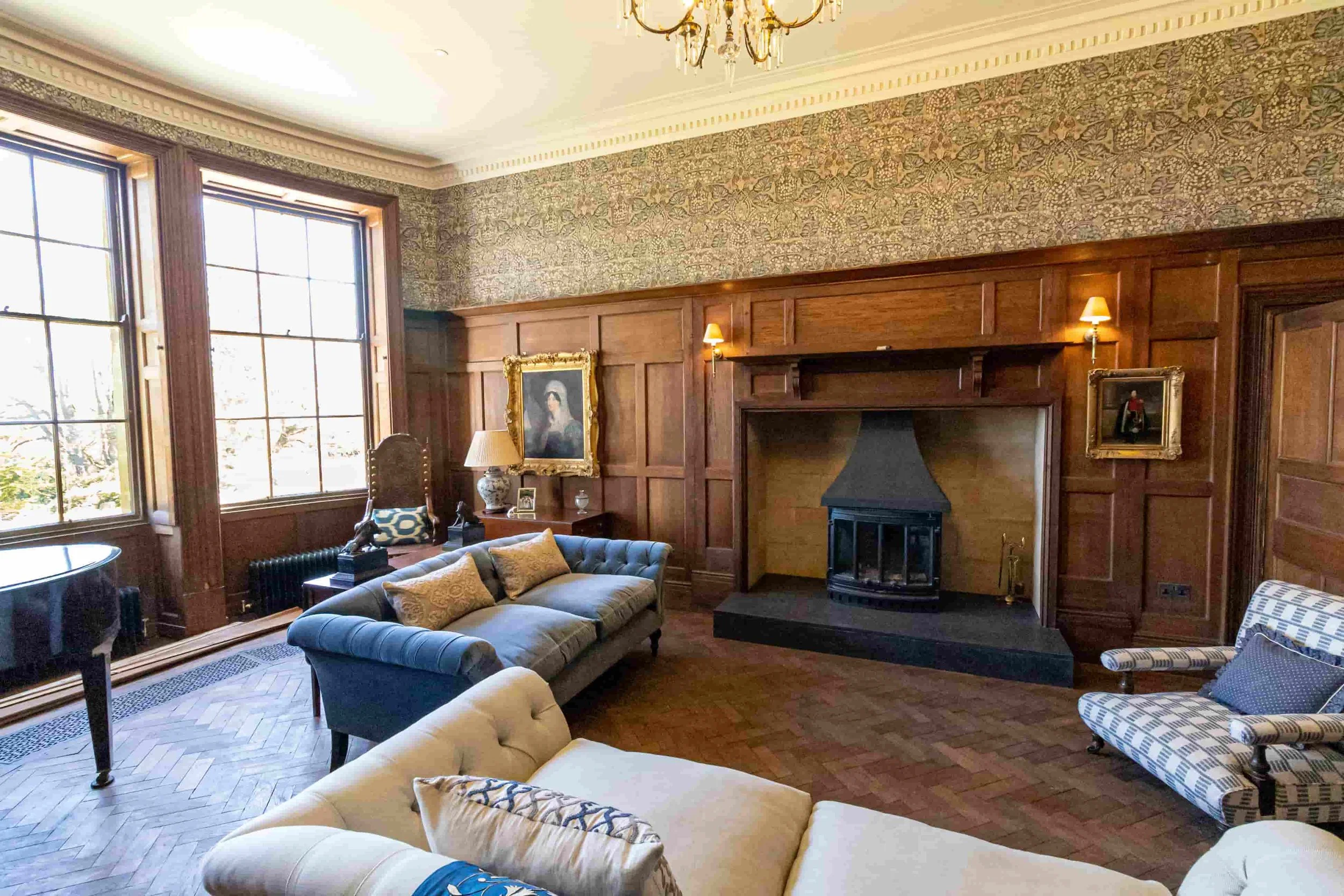 Elegant living room with large windows, wood-paneled walls, a fireplace, and vintage-style furniture, including sofas and armchairs, Victorian-style portraits, and decorative lamps.