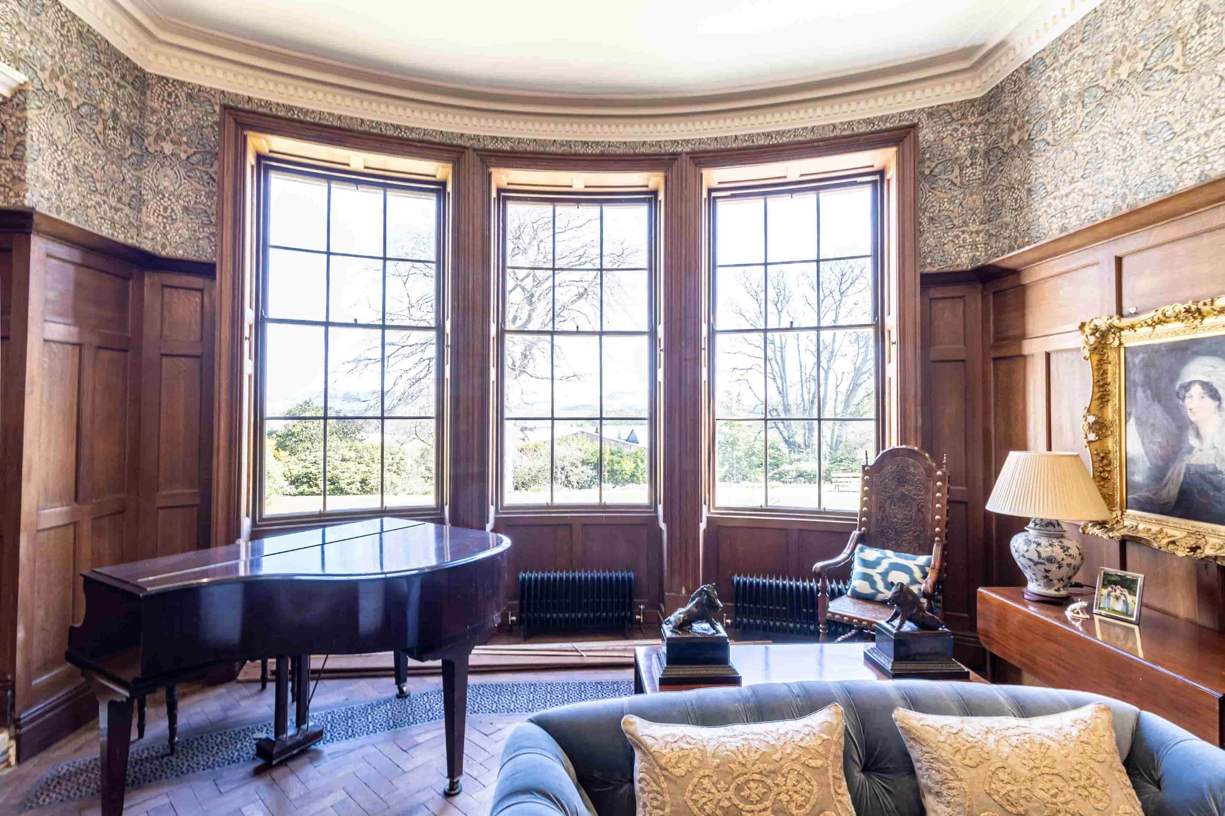 A luxurious living room with three large windows letting in natural light, a grand black piano, a wooden chair with a decorative pillow, a table with statues, a table lamp, a framed black and white portrait of a woman, and a sofa with pillows. The ro