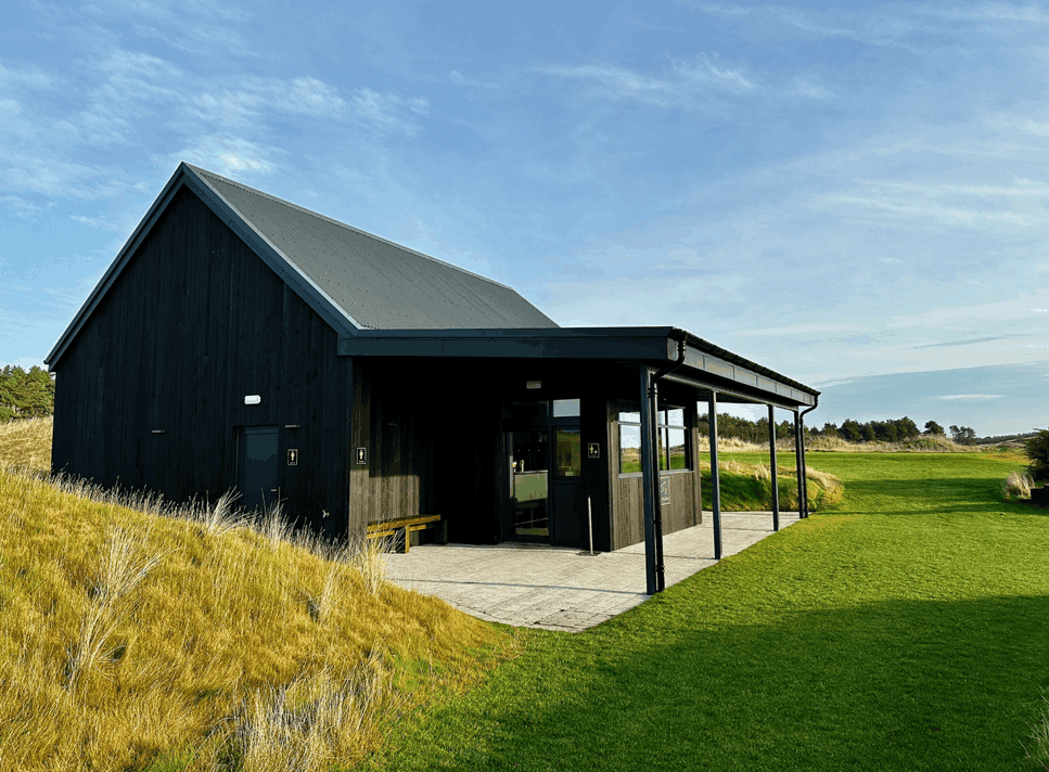A black wooden building with a slanted roof, situated on a grassy landscape with a patio area and a partly cloudy sky.