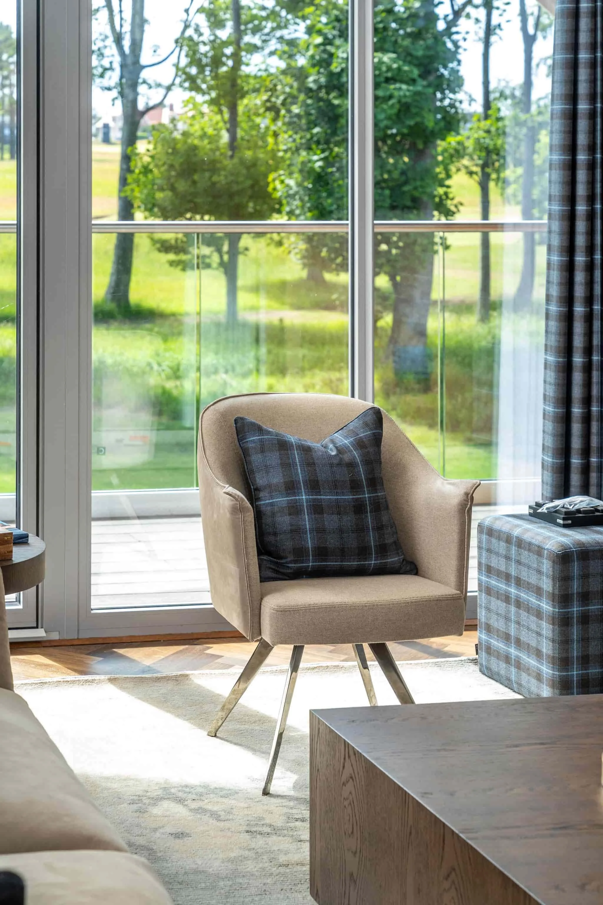 A cozy living room with a beige armchair, dark plaid pillow, wooden floor, and glass sliding doors leading to a balcony with a view of green trees and grass outside.