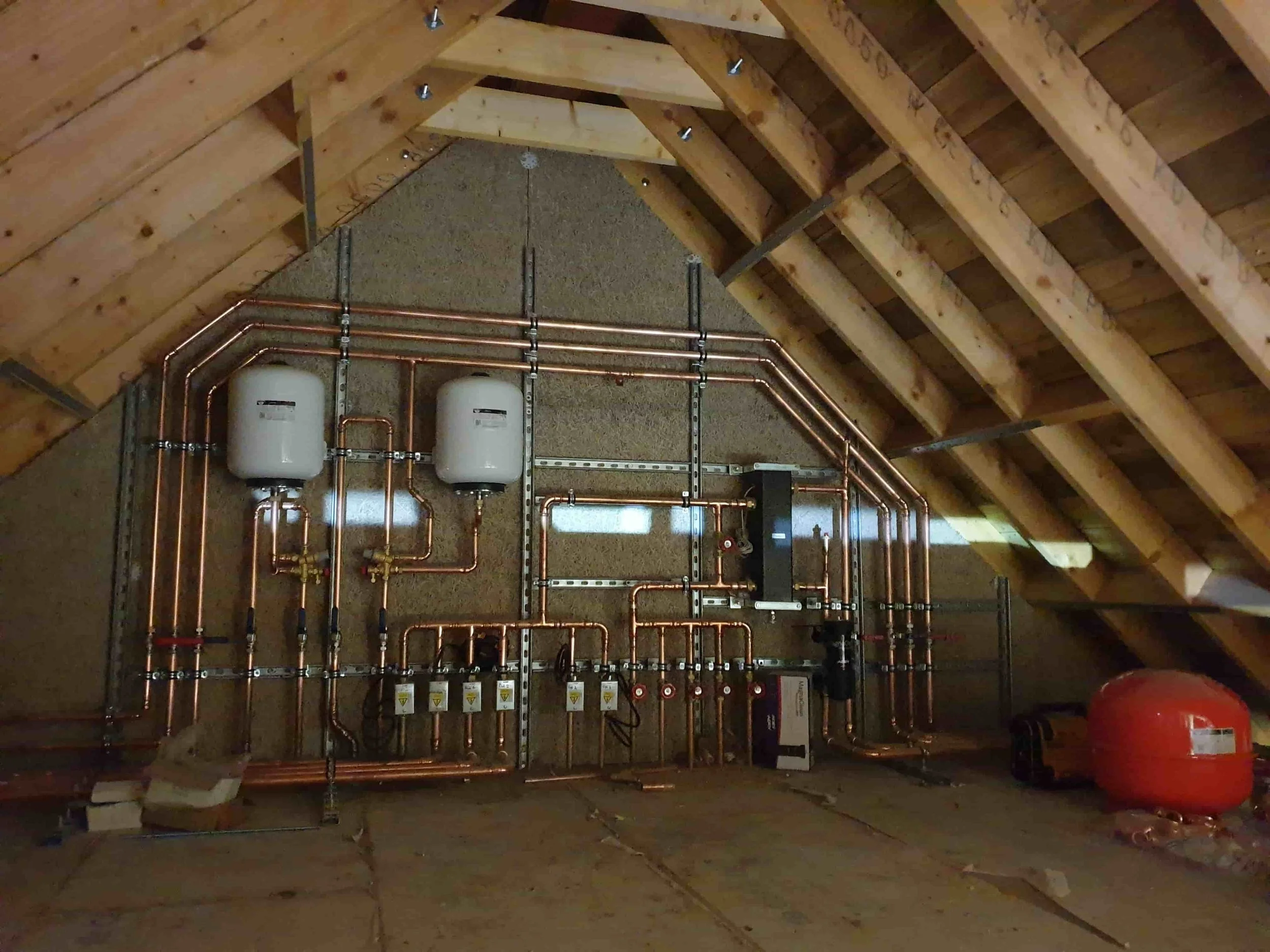 An unfinished attic with a copper piping system, two white tanks, and a red expansion tank installed on the back wall.