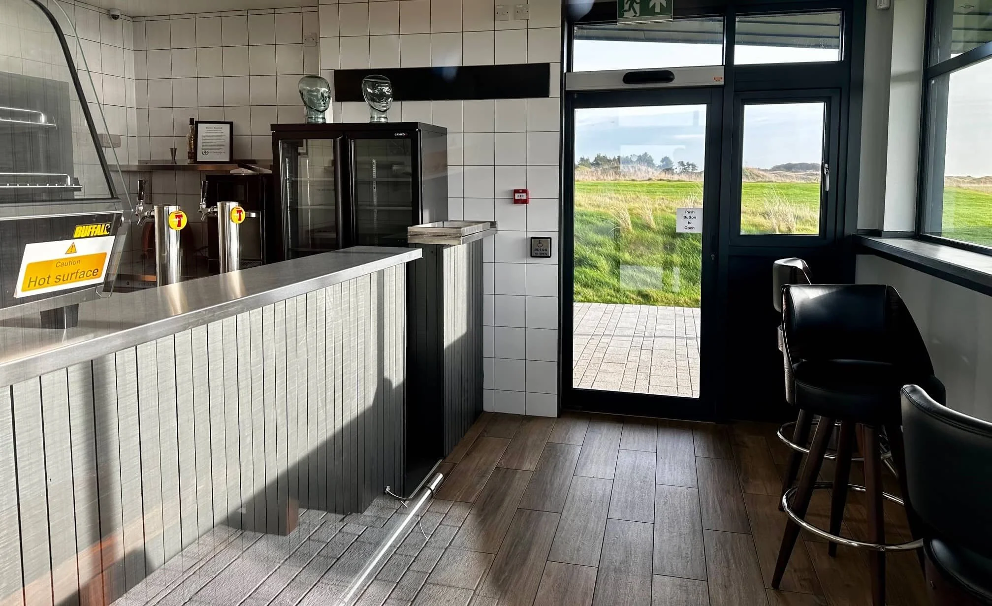 Interior of a beverage stand area with a counter, soda taps, a glass door, and a view of grassy outdoor landscape.