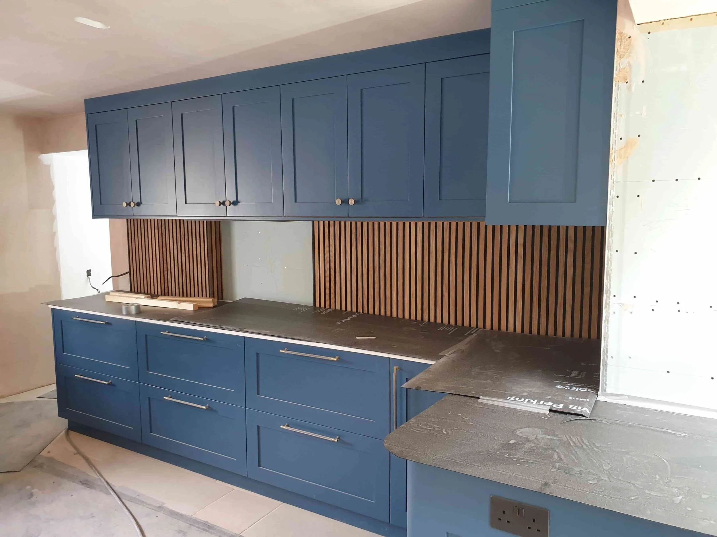 Blue kitchen cabinets with gold knobs, wooden slatted backsplash, and grey countertops under construction.