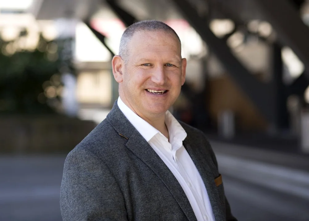 A smiling man in a white shirt and grey blazer standing outdoors with a blurred urban background.