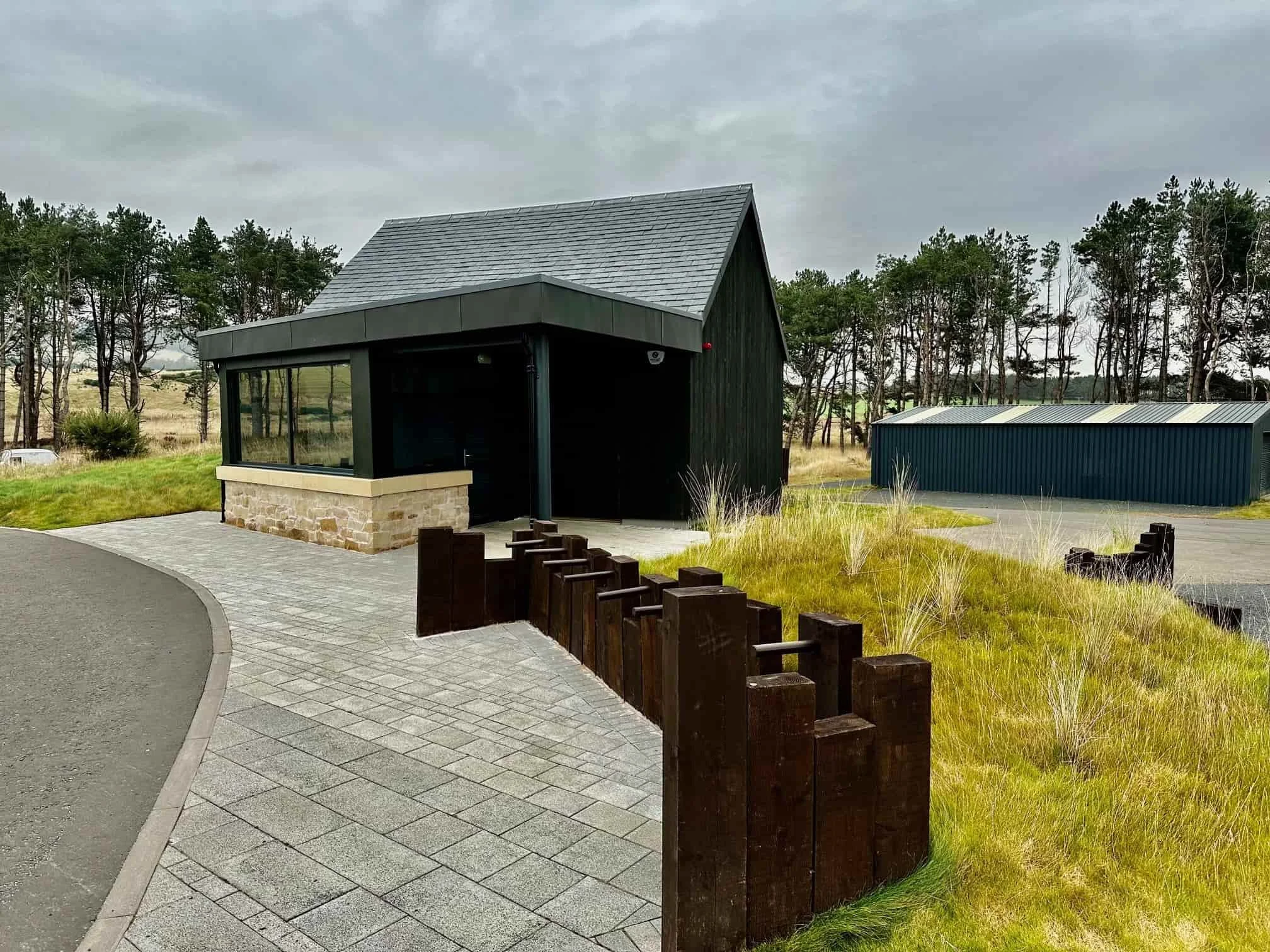 A small black building with large glass windows and a stone base, located in a natural setting with trees and grass. A paved walkway leads to the building, with a wooden barrier on one side. There is a large blue metal storage shed in the background.
