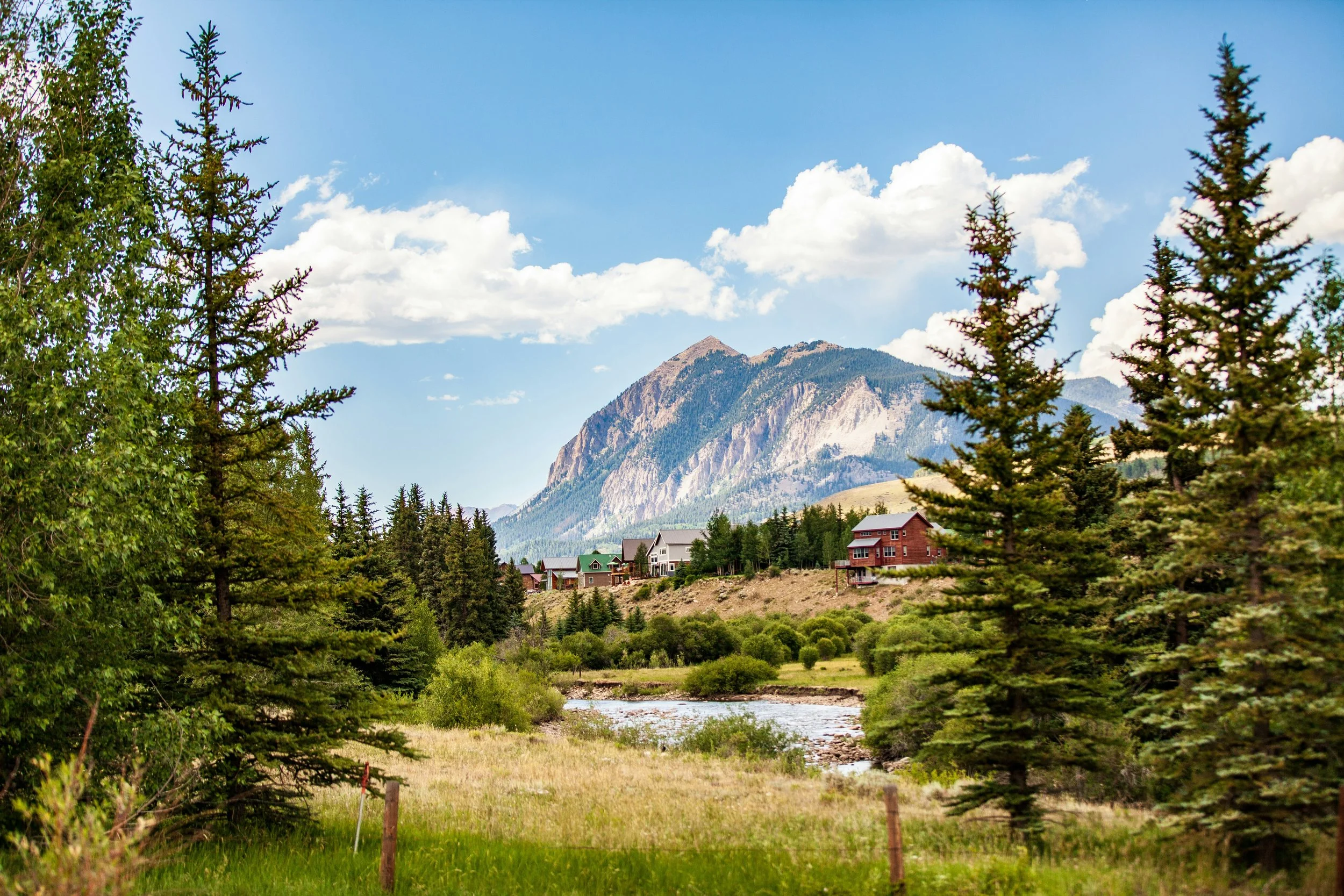 A scenic mountain landscape with a small river in the foreground, surrounded by lush trees and houses, with mountain peaks and a partly cloudy sky in the background.
