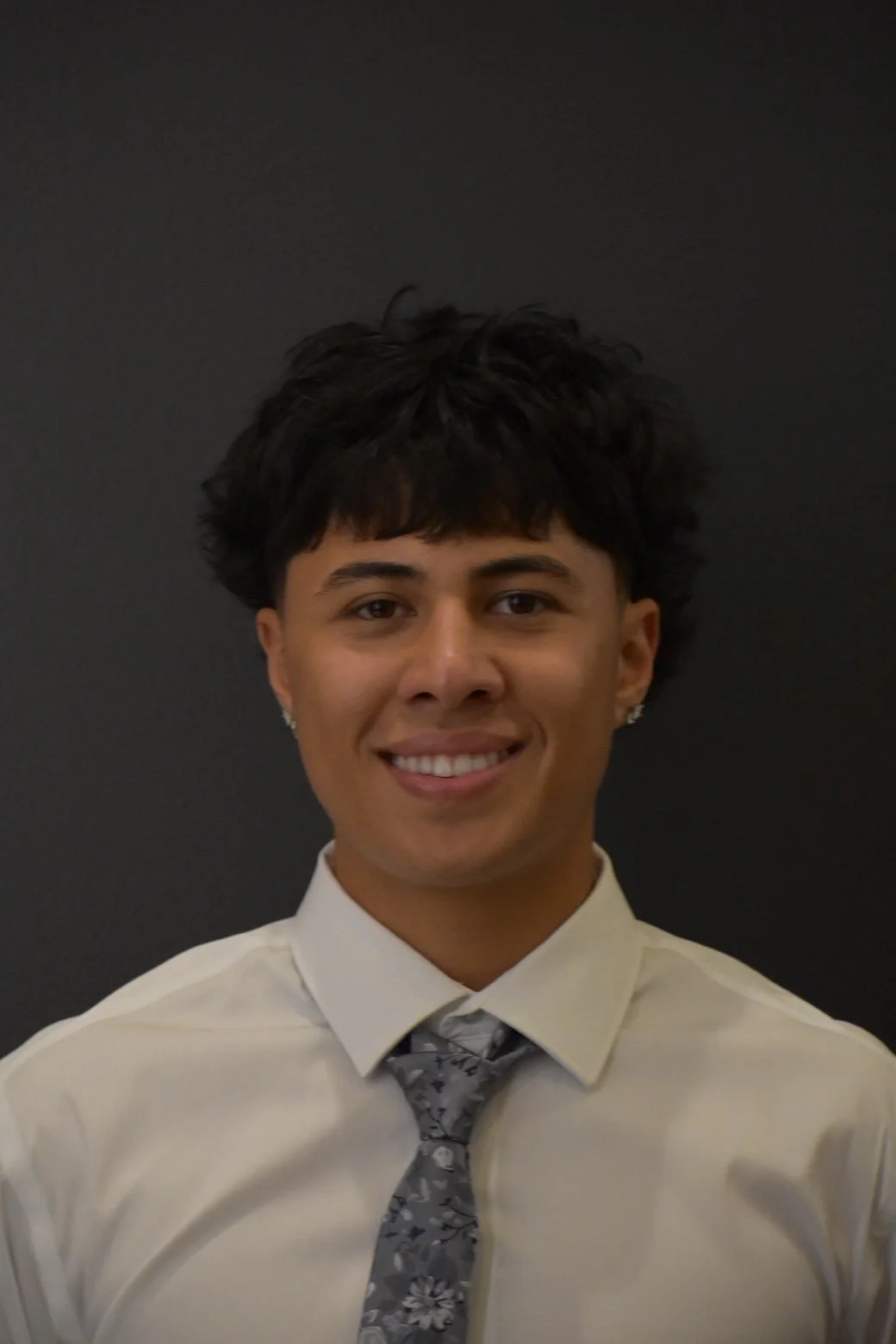 A young woman with short, curly dark hair dressed in a white dress shirt and a floral gray tie, standing against a dark background and smiling at the camera.