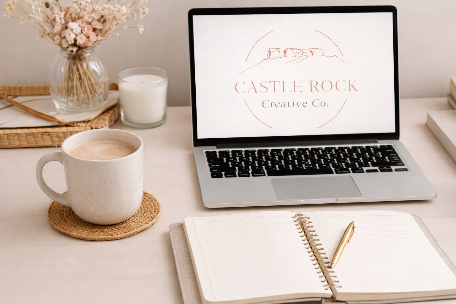 A workspace with a laptop displaying 'Castle Rock Creative Co.' logo, a white mug of coffee on a woven coaster, an open notebook with a gold pen, a glass of candle, and a vase with pink flowers.