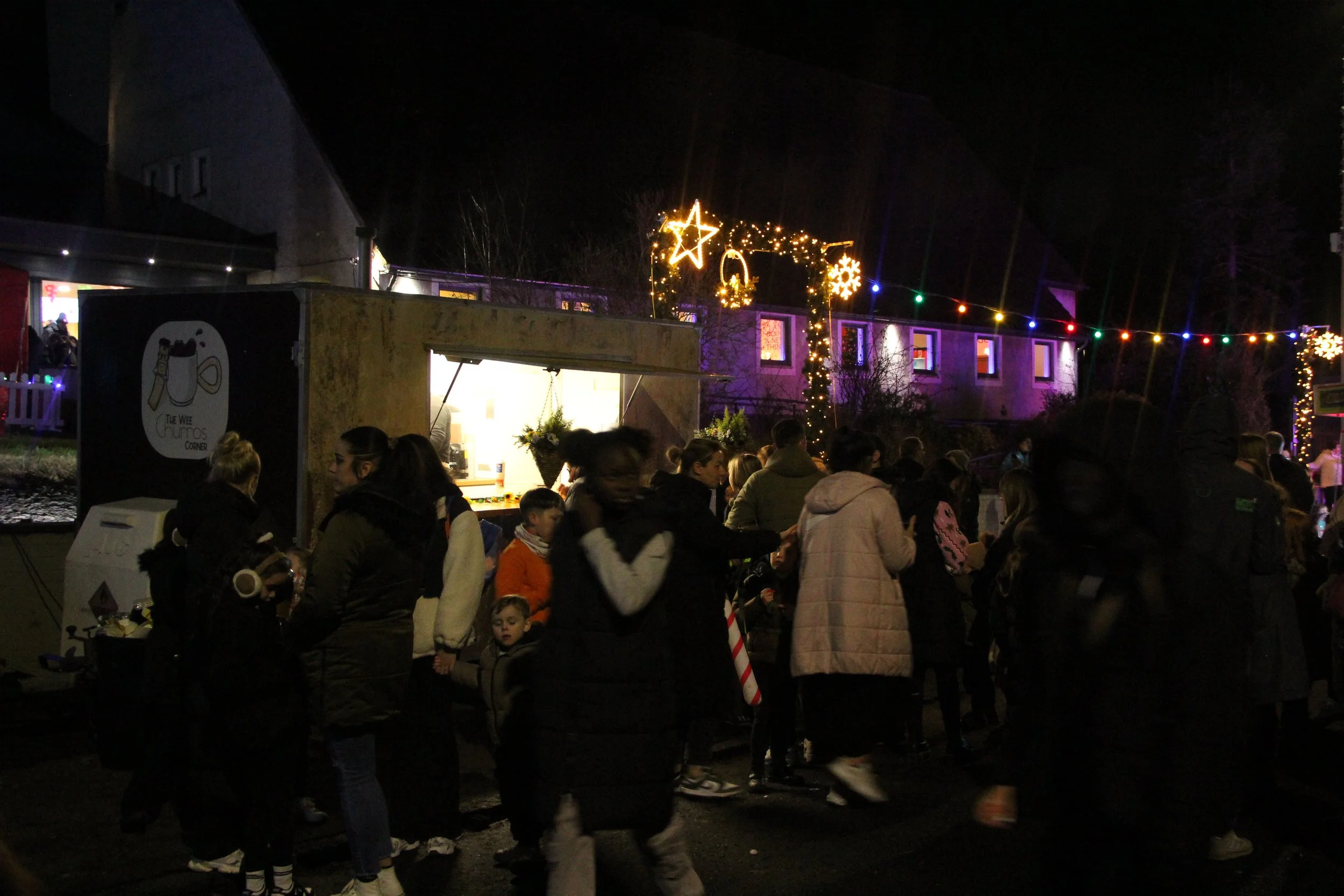 Night scene of a festive outdoor market with people gathered around a food stall, decorated with Christmas lights, stars, and snowflakes, with a house lit up in the background.