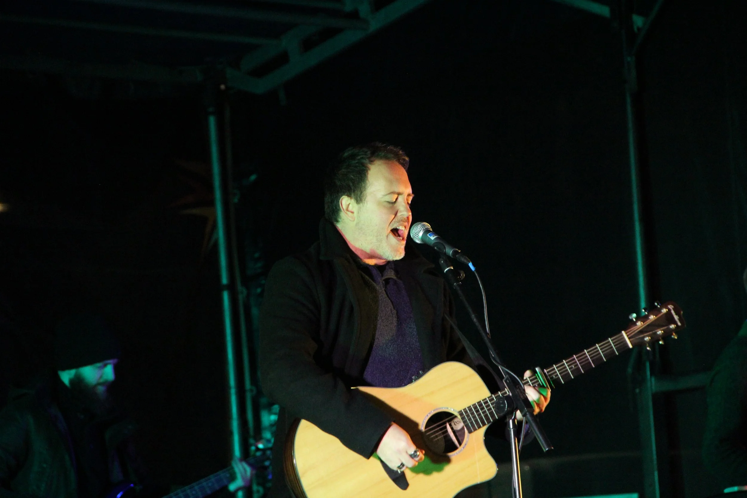 A man singing into a microphone while playing an acoustic guitar on stage.
