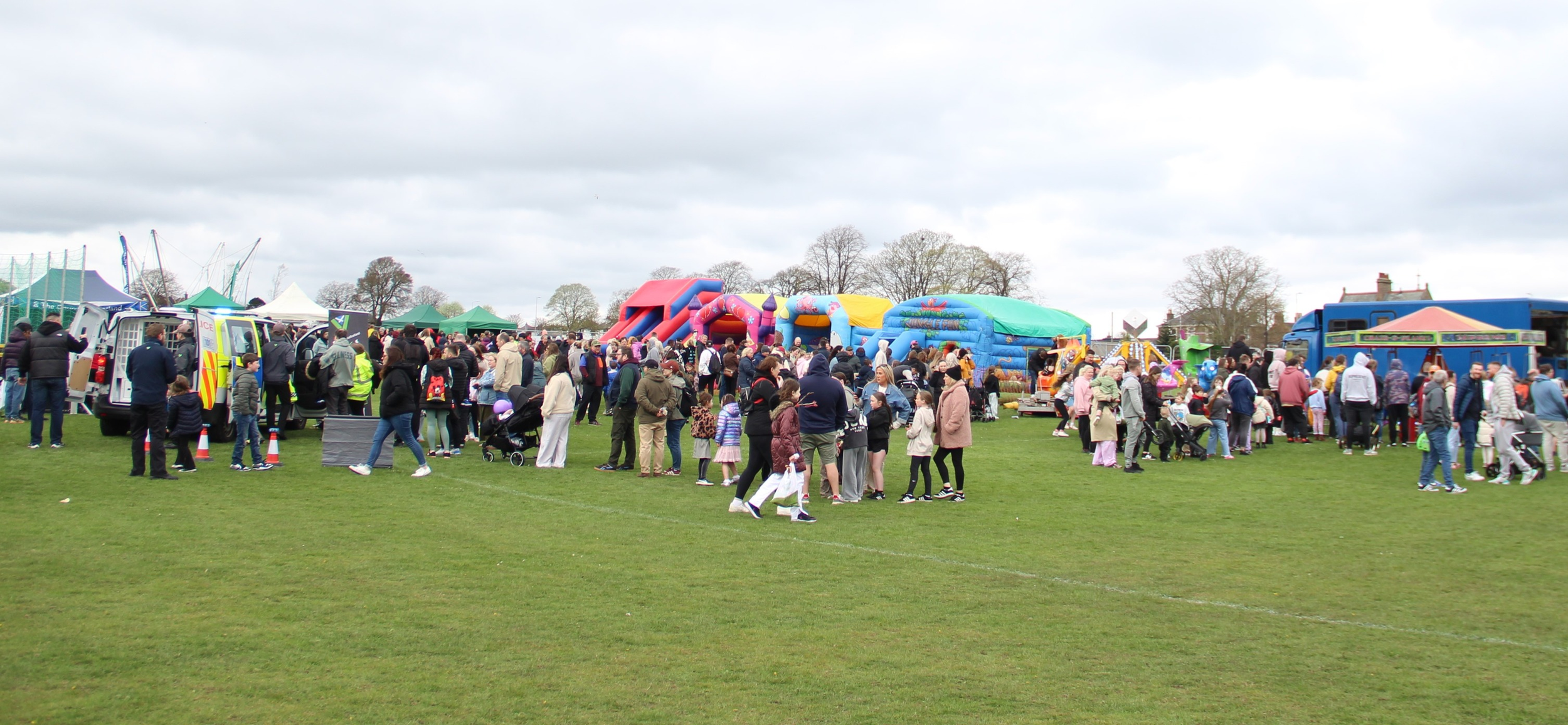 Crowd of people at an outdoor fair with adults, children, and inflatable bounce houses on a grassy field under cloudy sky.