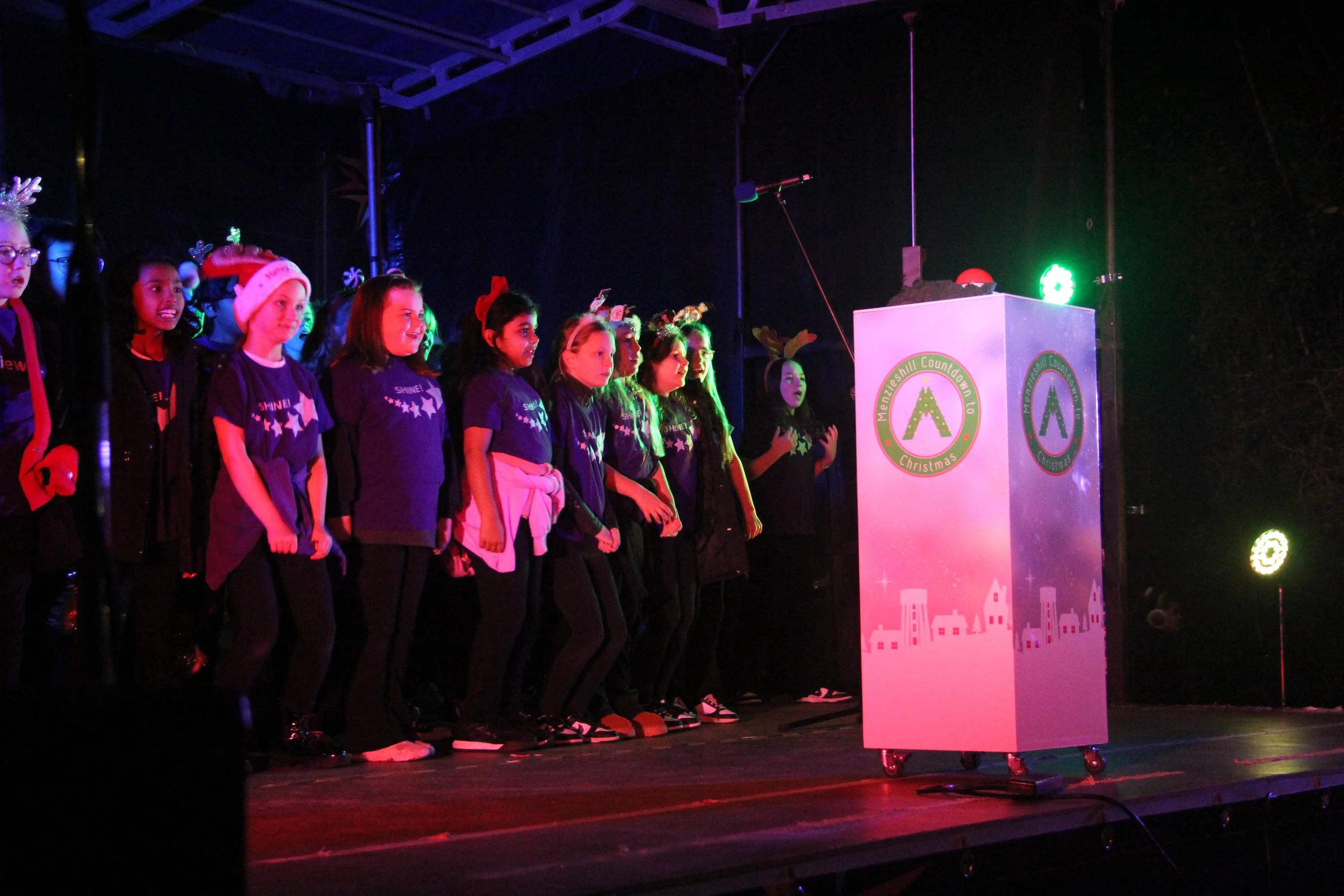 Children on stage at Christmas event, some wearing holiday hats and reindeer antlers, standing in a line facing a microphone and a decorated podium.