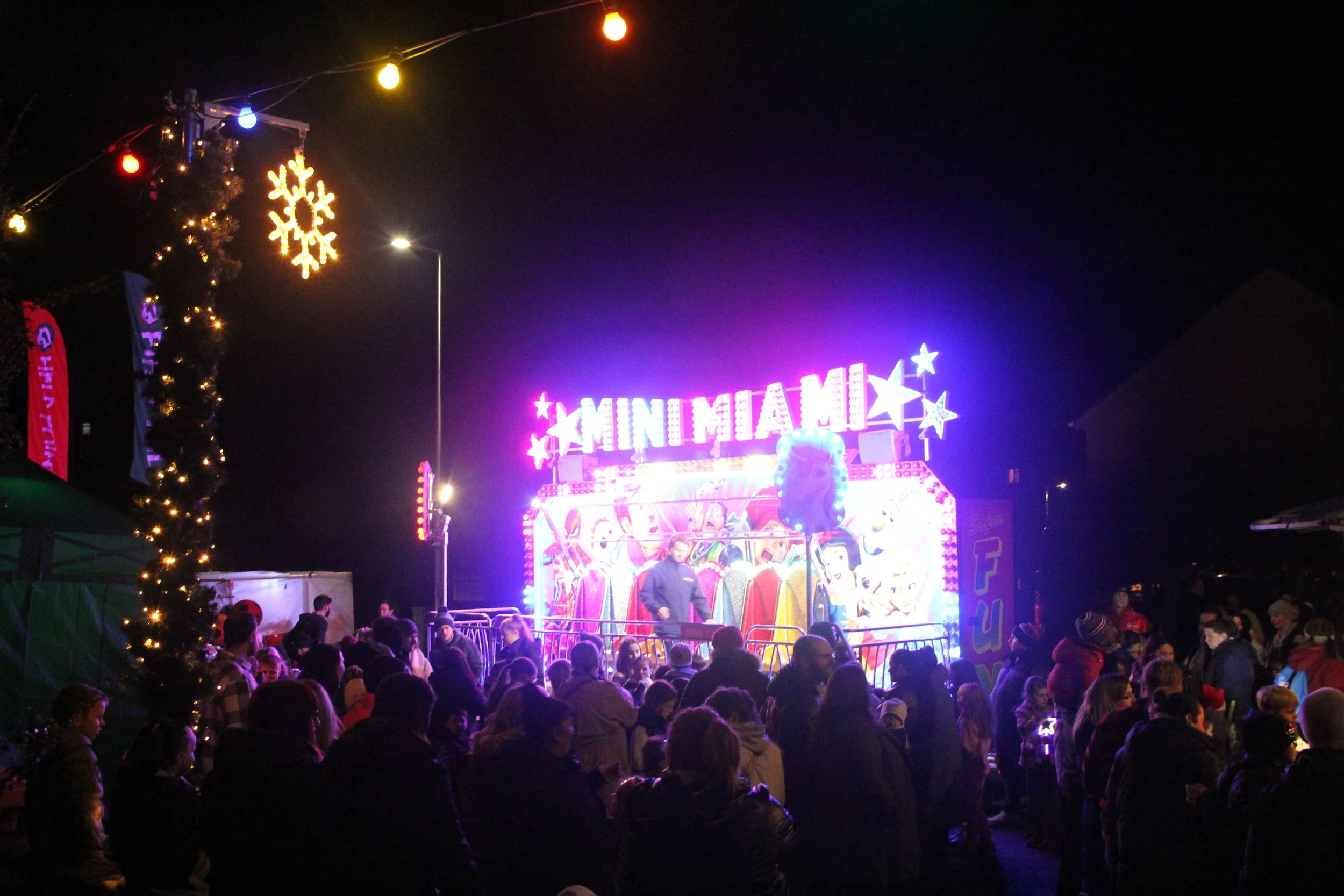 A lively outdoor carnival or fair at night with a brightly lit amusement ride called 'Minimiam' featuring animated characters and pink, purple, and yellow lights. A crowd of adults and children gathers around, enjoying the festive atmosphere. Holiday decorations like colorful string lights and a snowflake ornament are visible.