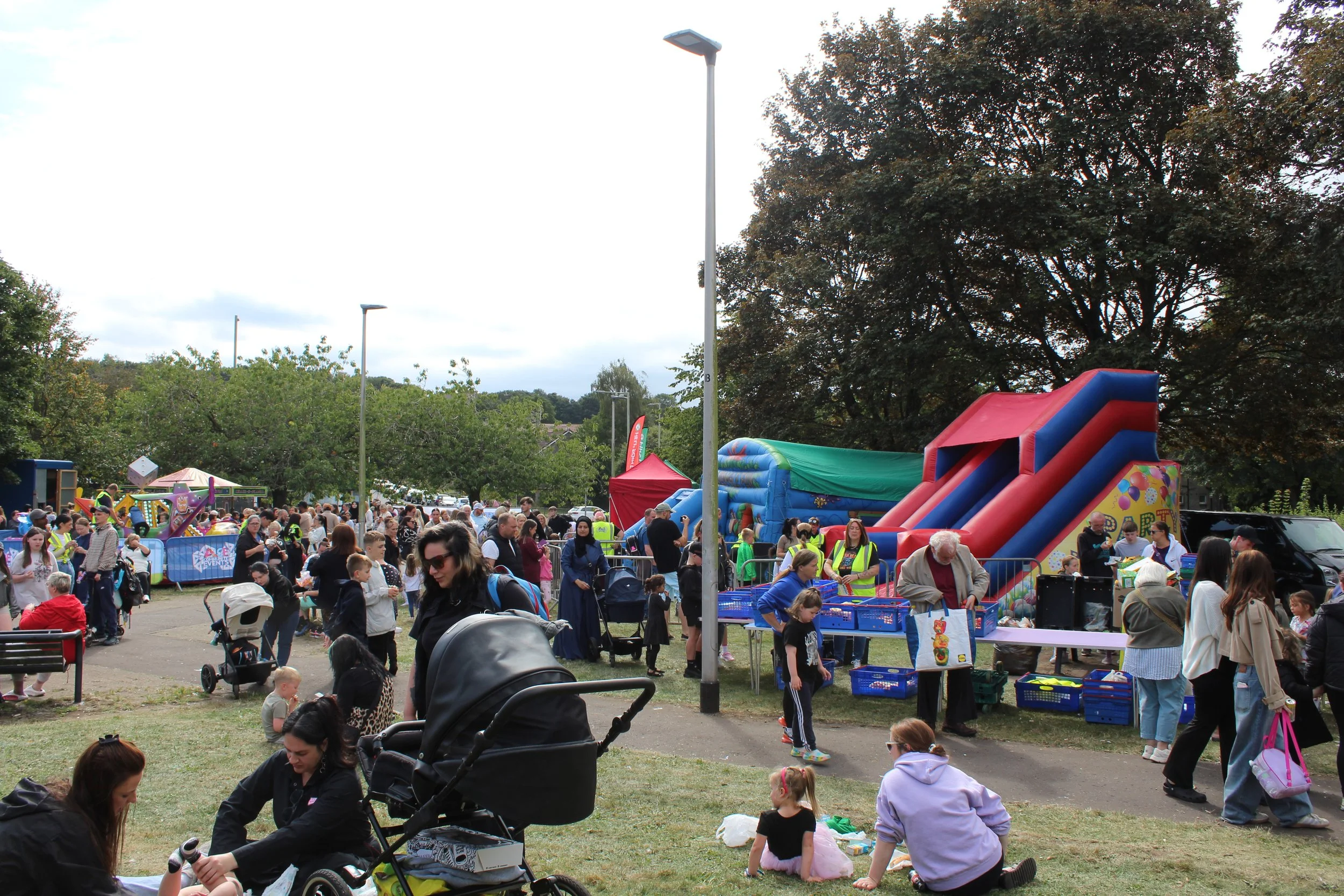 A crowded outdoor fair with people, children, and inflatable slides, with trees and cloudy sky in the background.