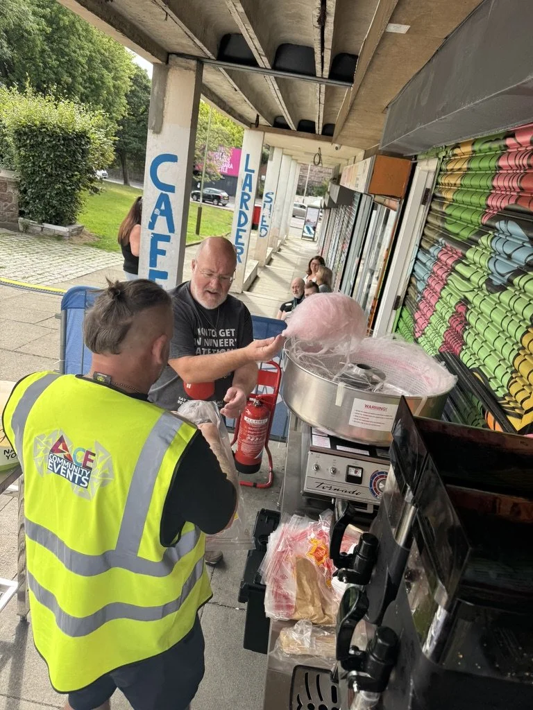 Man in black shirt handing cotton candy to another person in a yellow vest at an outdoor fair or event stand with colorful rolled paper supplies on the right and a fire extinguisher nearby.