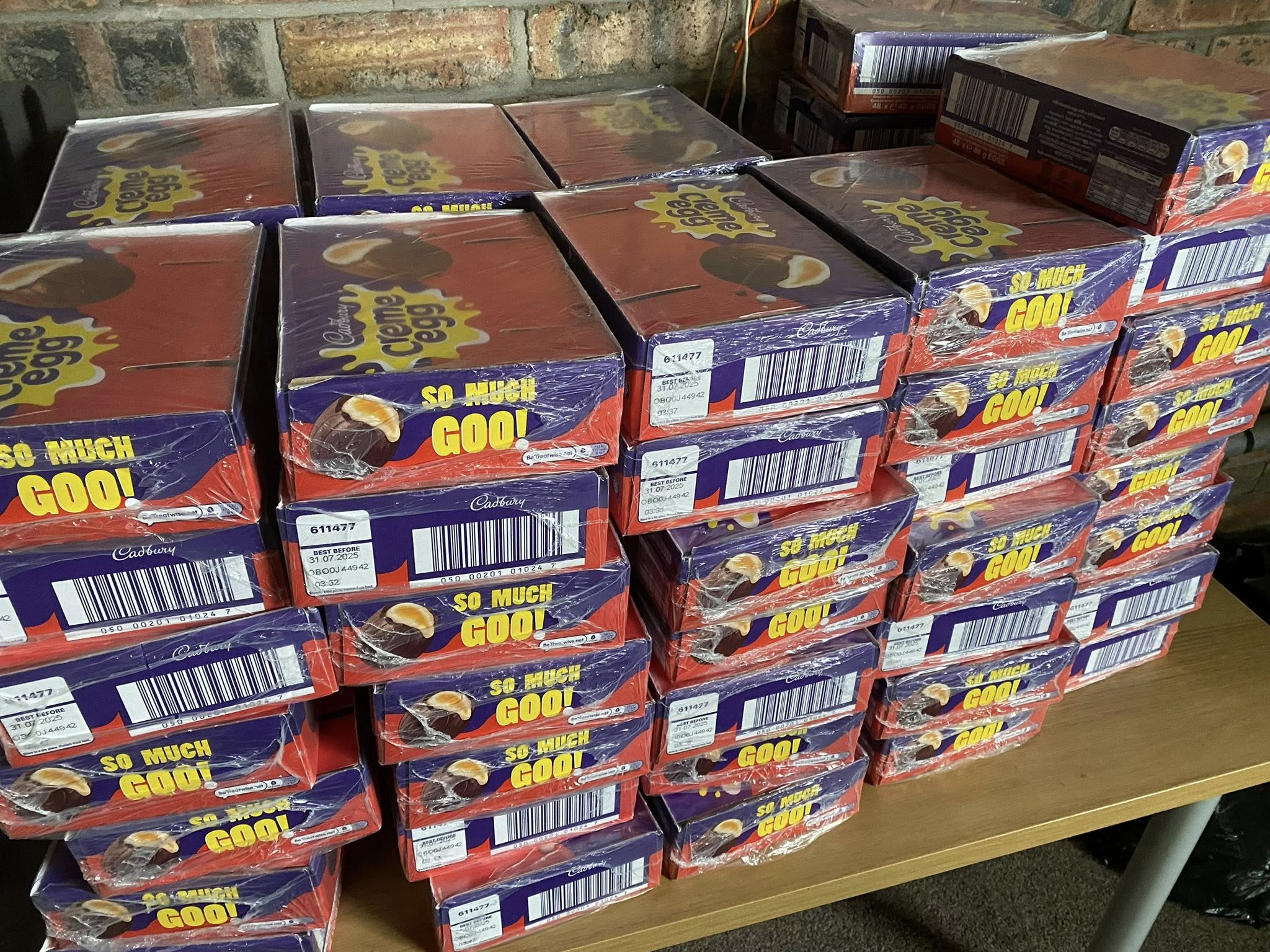 Several rows of Cadbury chocolate egg boxes stacked on a wooden table against a brick wall, with a colorful purple and red design and the phrase 'SO MUCH GOO!' on the packaging.