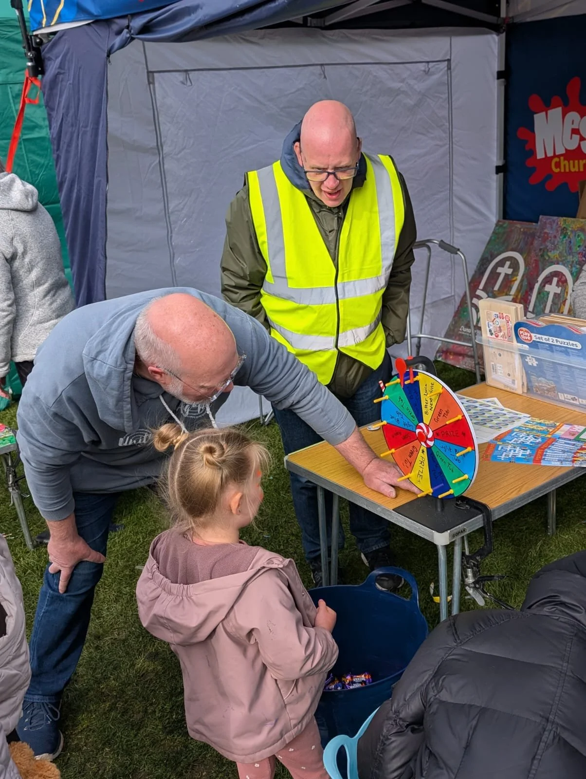 A man in a gray hoodie spins a colorful prize wheel while a young girl and an older man watch. The scene is outdoors with a blue tent in the background, and there are various items on a table nearby.