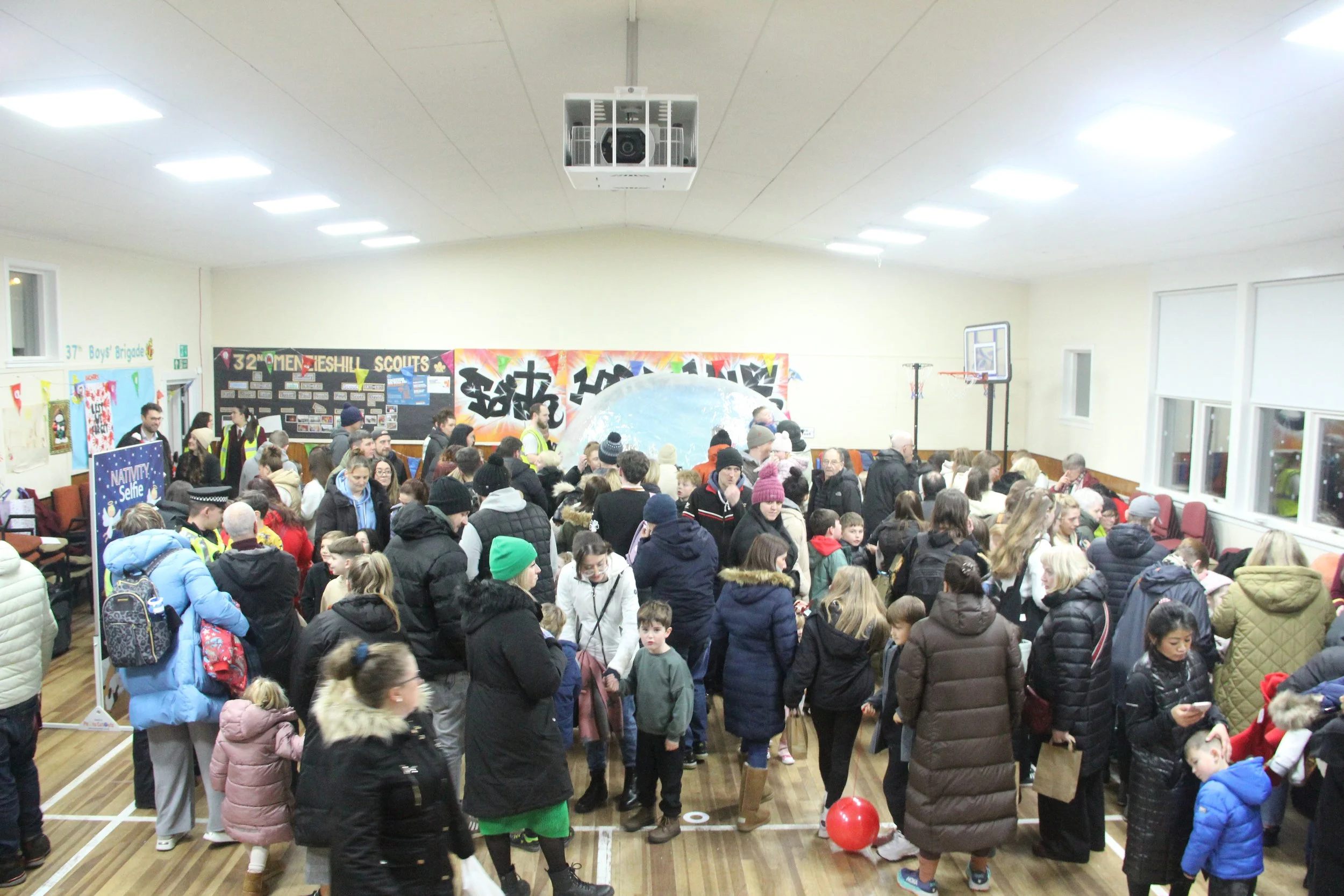 A crowded community event in a gymnasium with many children and adults, colorful decorations, posters, and a large globe and a basketball hoop in the background.