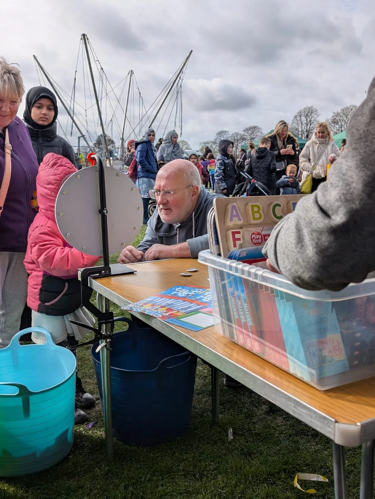 A man and children at a game booth outdoors at a fair or festival, with people and sailboats in the background under cloudy skies.