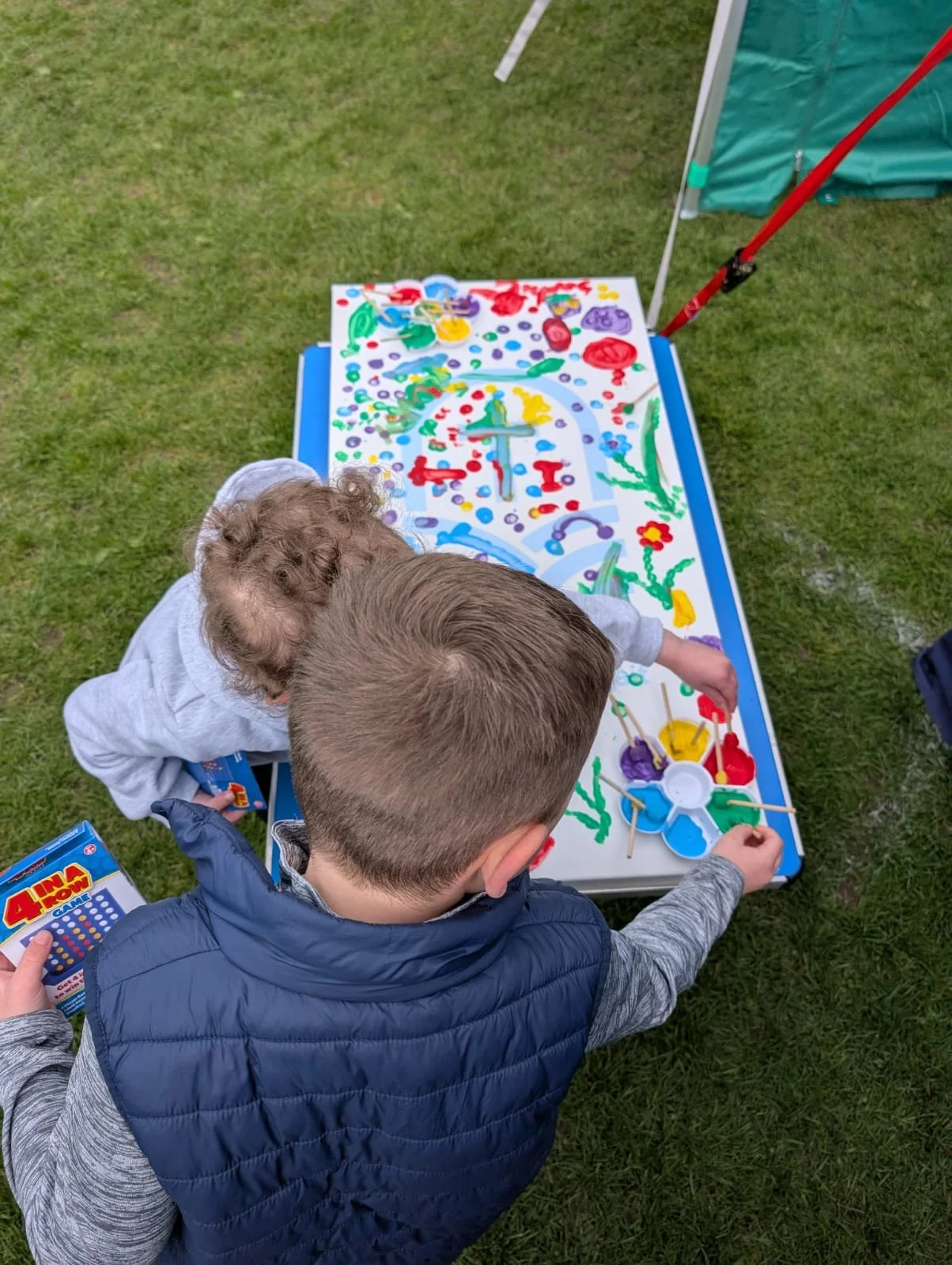 Two children painting a colorful picture on a white desk outdoors, with a tent nearby, on a grassy field.
