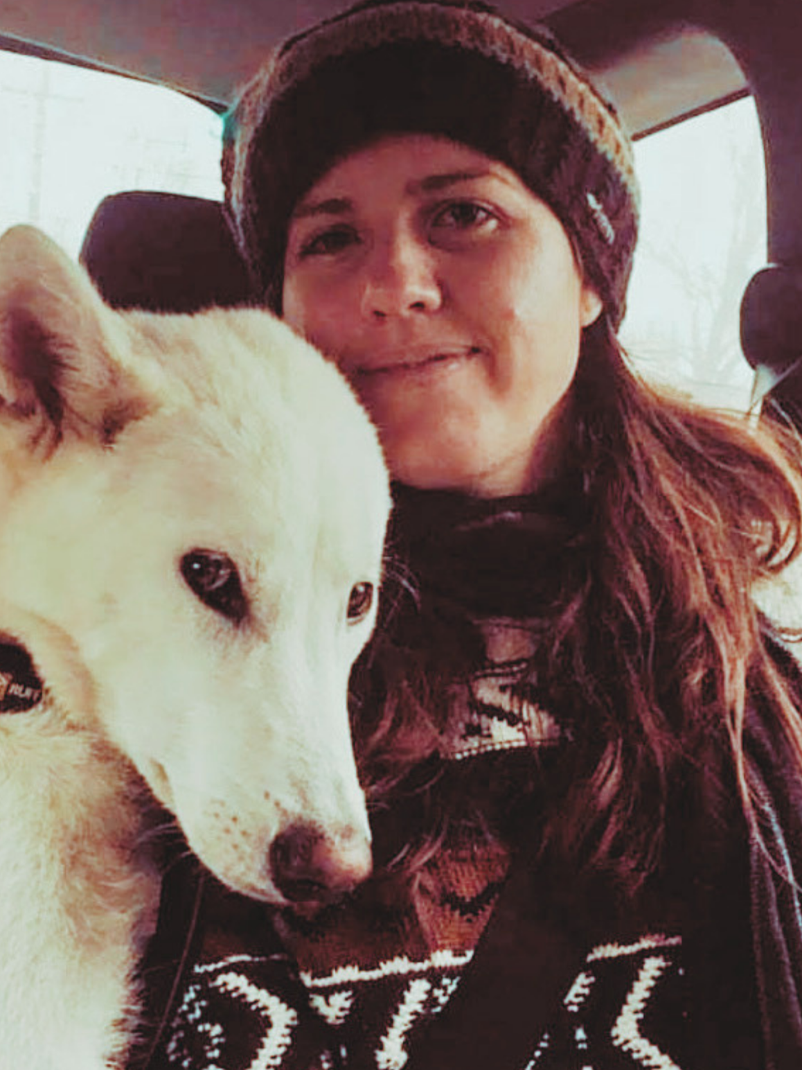 Picture of white woman with long brown hair wearing a winter sweater and beanie sitting in car smiling sitting next to a white german shepard dog.