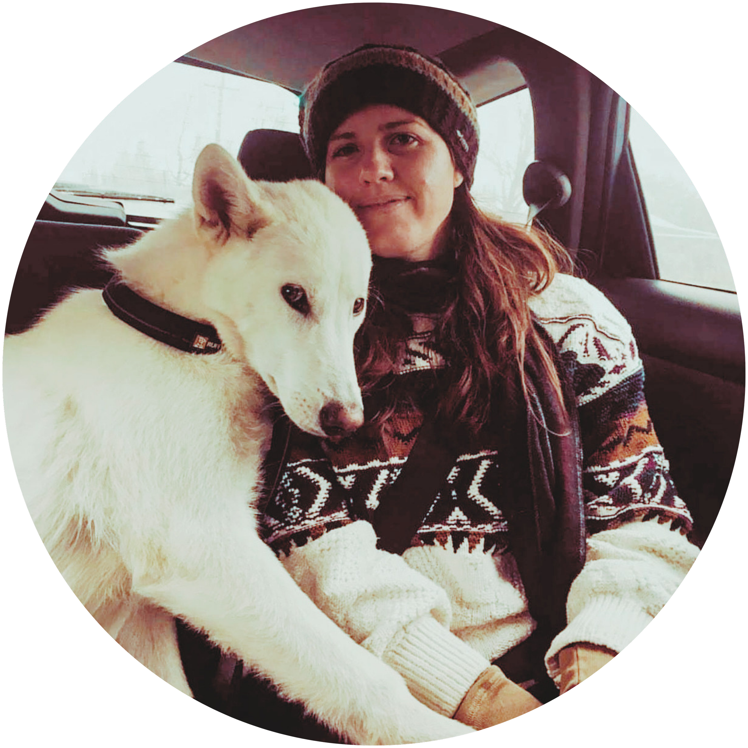 Picture of white woman with long brown hair wearing a winter sweater and beanie sitting in car smiling sitting next to a white german shepard dog.