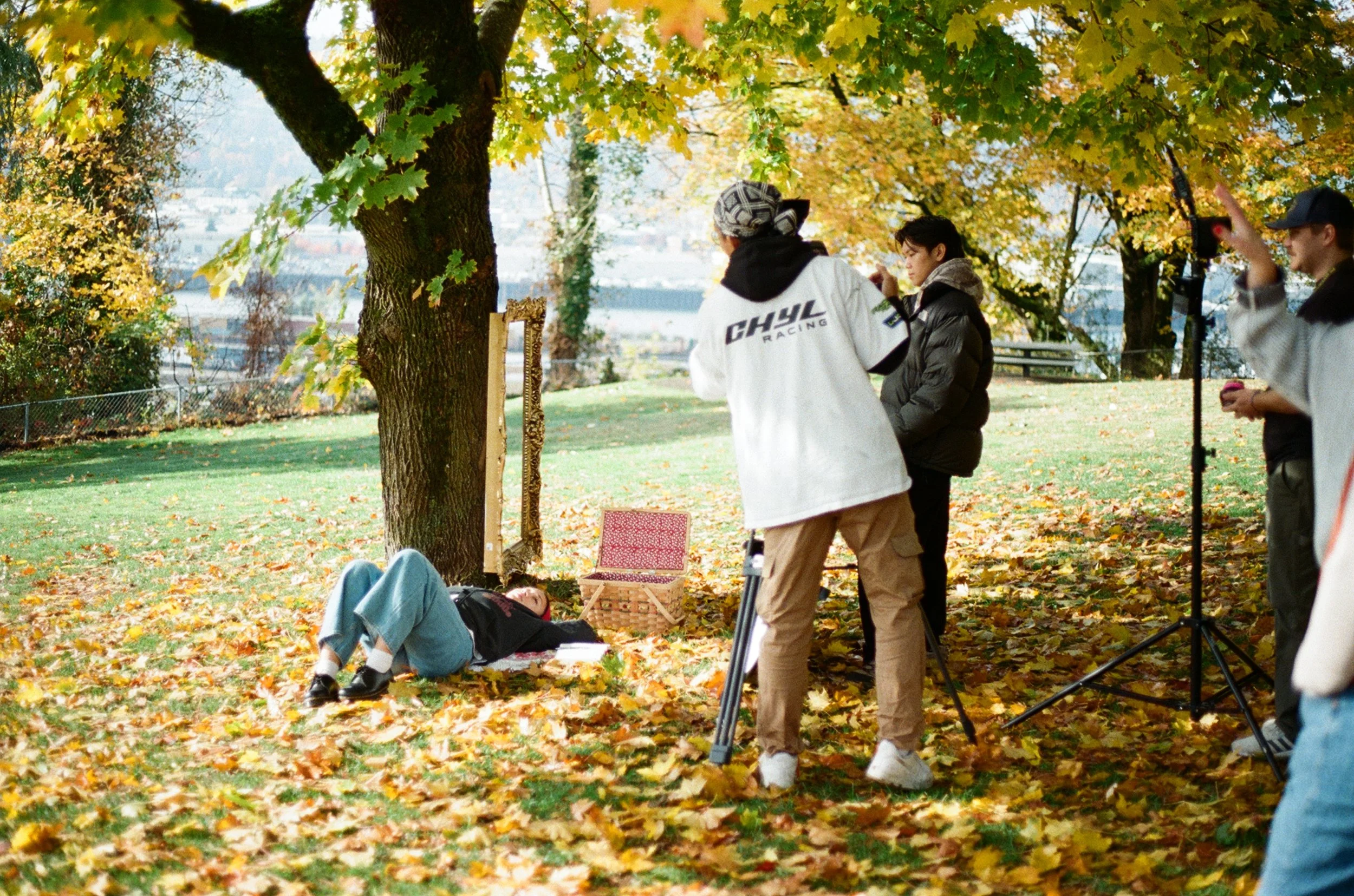 Film crew filming a scene outdoors in a park during fall, with autumn leaves on the ground and trees with yellow and orange leaves, a person lying on a blanket, and various crew members setting up equipment.