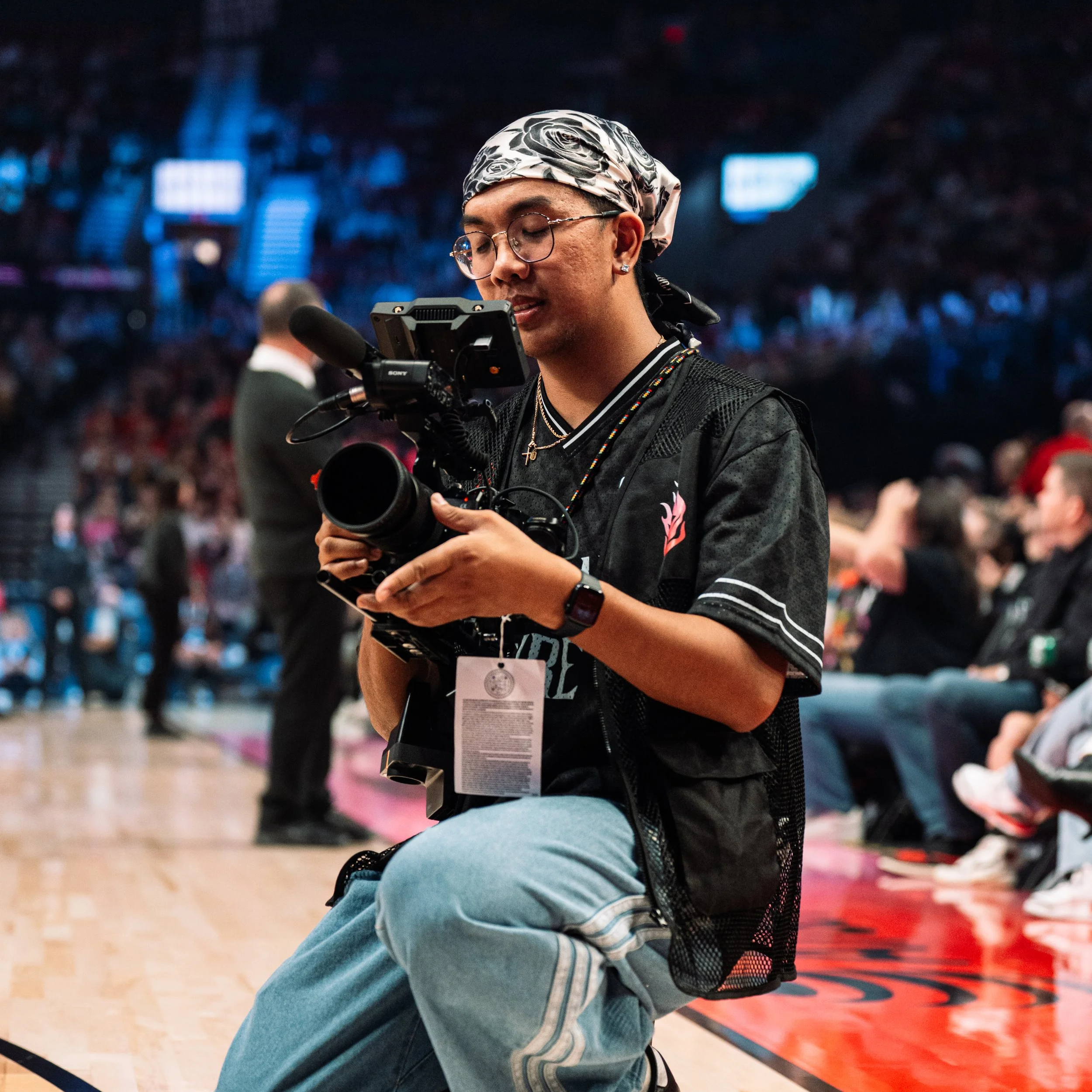 A person with glasses and a patterned headscarf, sitting on the floor in a sports arena, filming or taking pictures with a camera. The person is wearing a black shirt and jeans. There are blurred spectators in the background.
