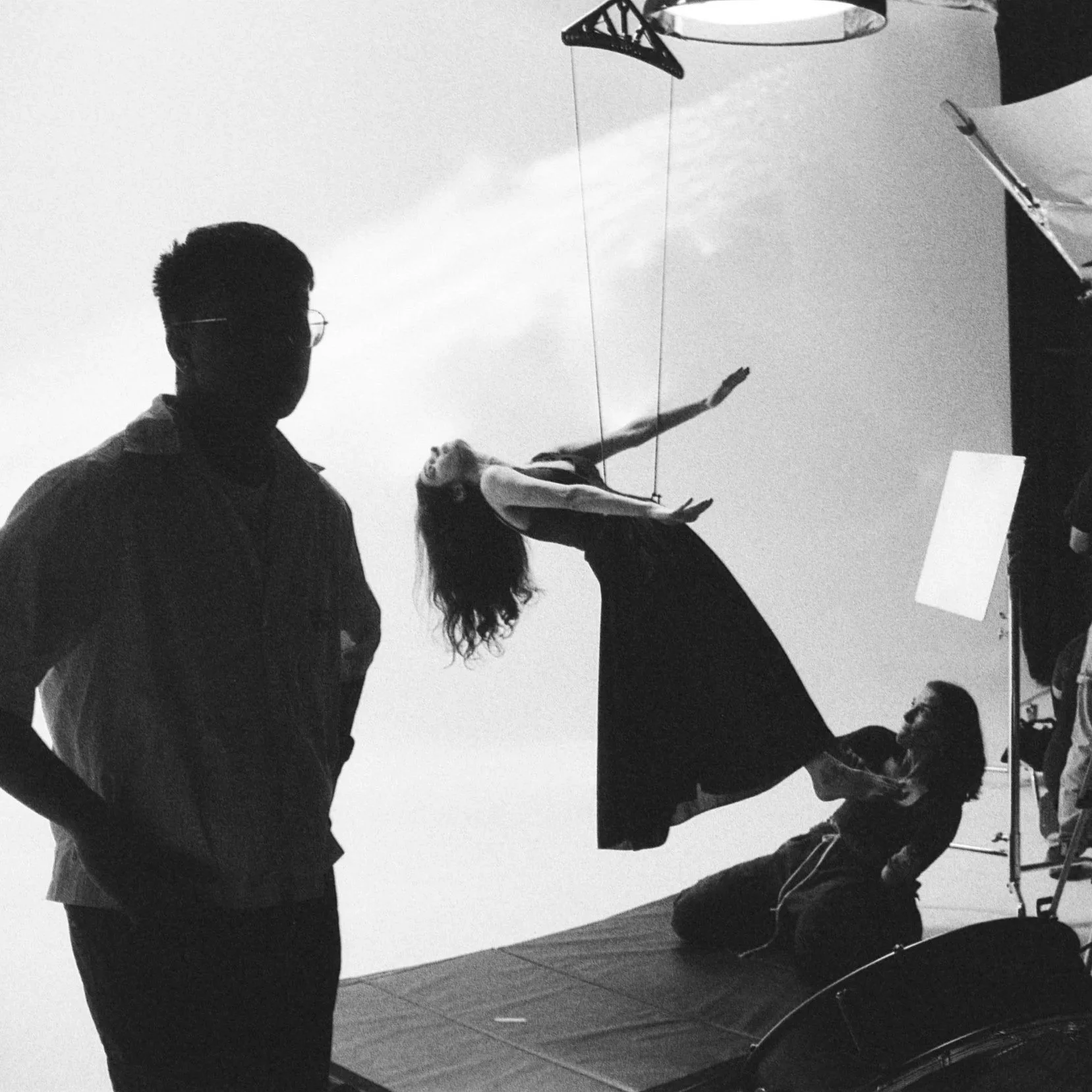 People on a photography or film set, with one person holding a large reflector and another person adjusting a cloth or backdrop, all silhouetted against a light background.