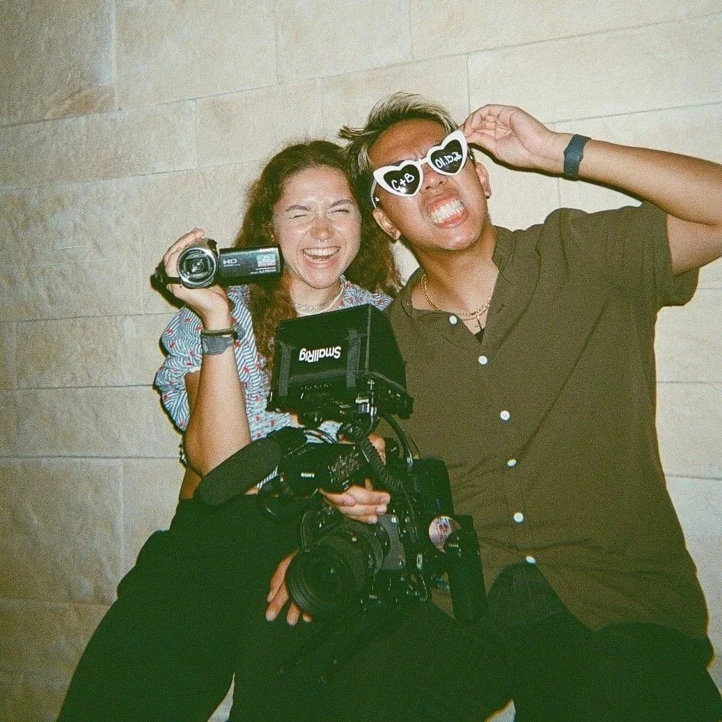 Two young people, a woman and a man, are having fun and taking photos together. They are standing against a light-colored wall, with the woman holding a video camera and a large professional camera hanging from her neck. The man is wearing dark sungl