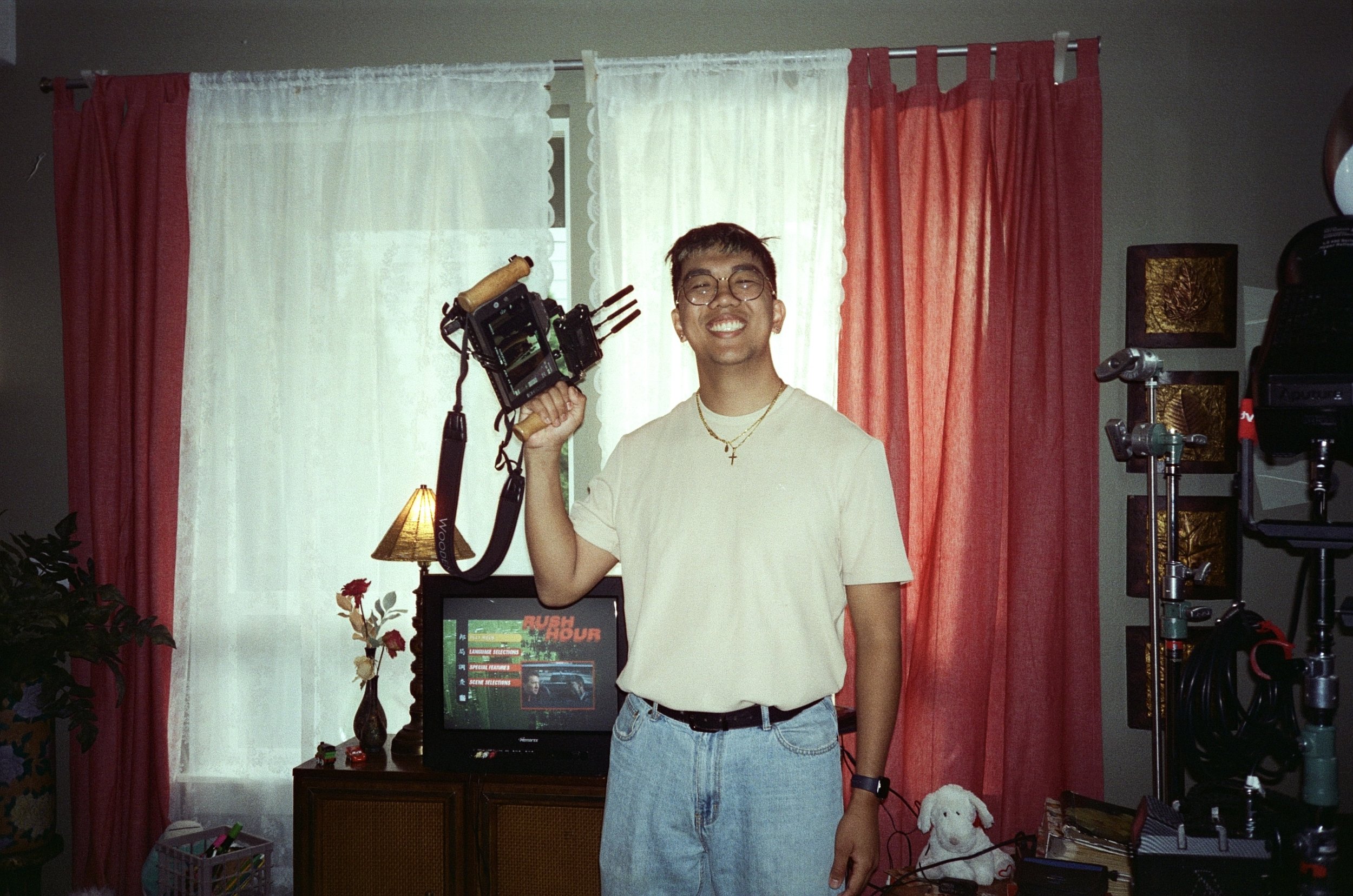 Young man smiling, wearing glasses, with a camera stabilizer on his shoulder, standing in a room with red and white curtains, a TV on a wooden cabinet, and various lights and equipment.