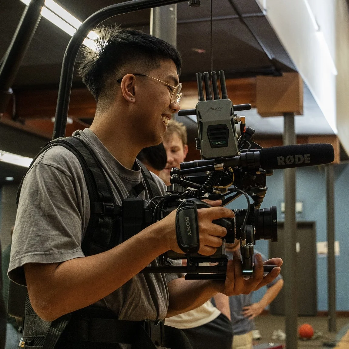 A man operating professional film equipment indoors, with people in the background.
