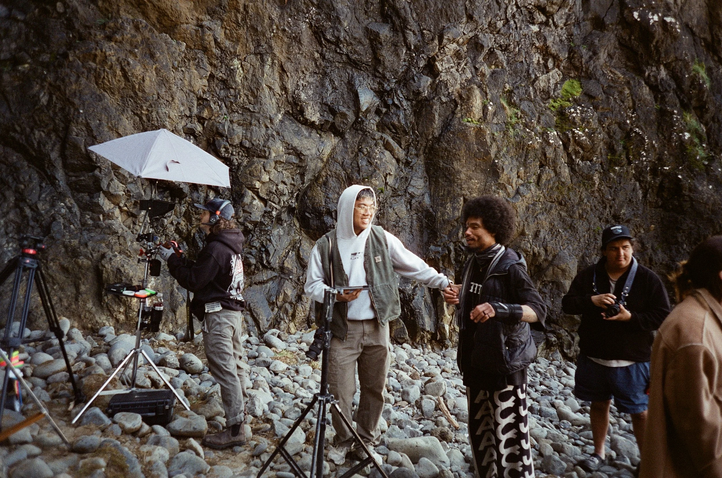 Group of people standing on rocky terrain next to a large rock face with filming equipment around, some holding cameras and others engaging in conversation, in an outdoor setting.