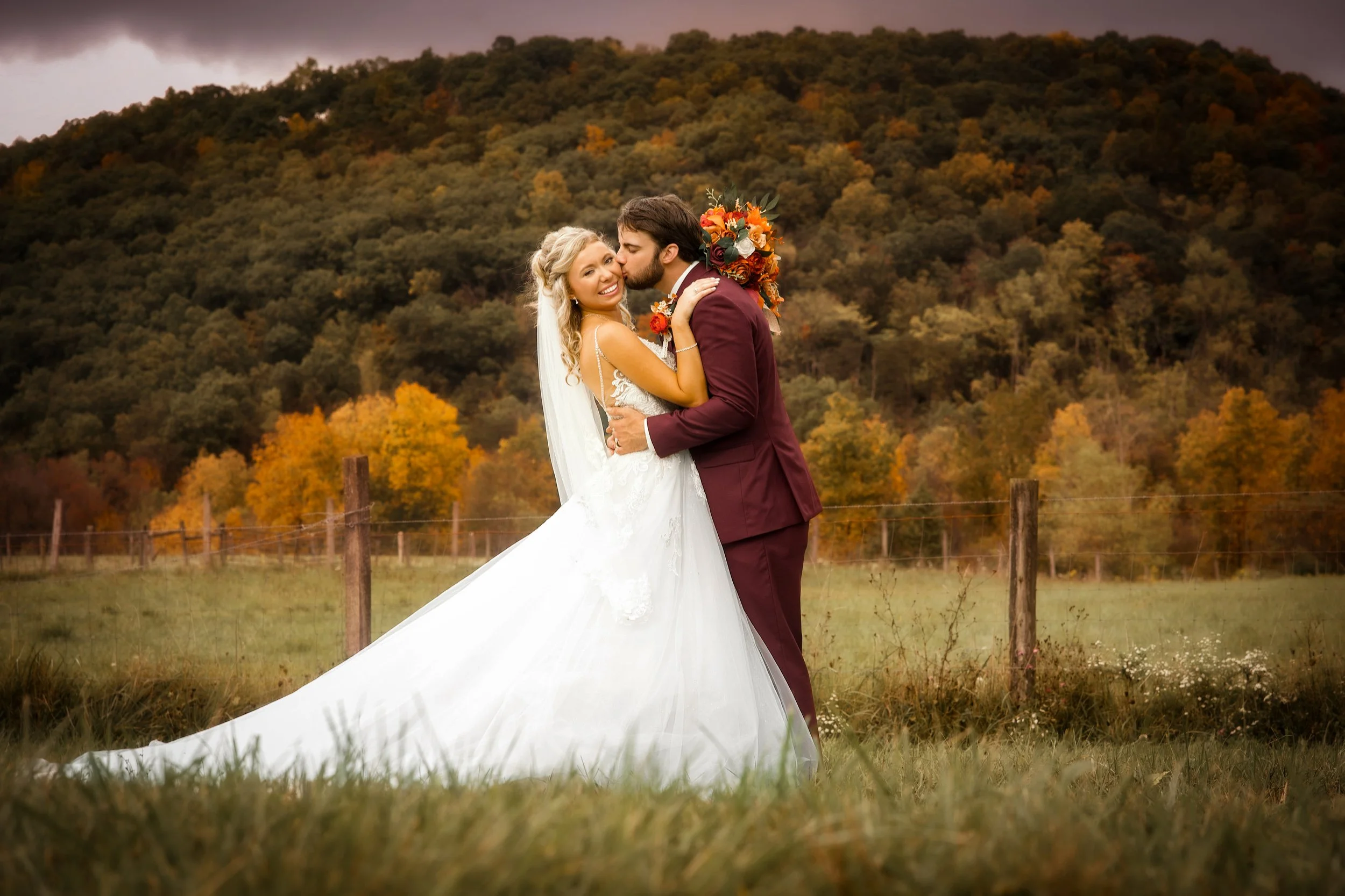 A bride and groom standing together outdoors in a grassy field with autumn trees and a hill in the background. The groom is kissing the bride on the cheek, and the bride is smiling.
